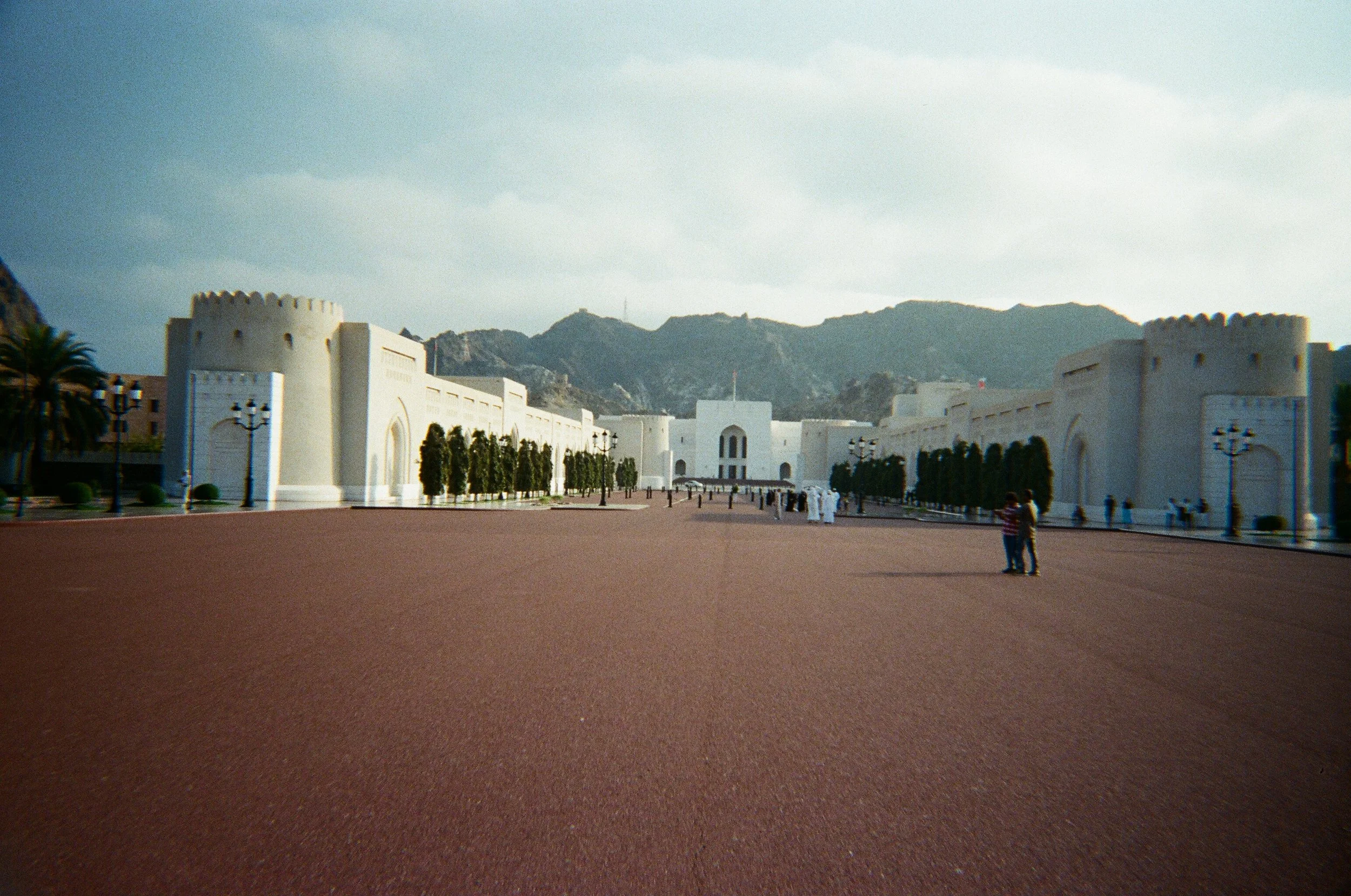 Una grande fortezza bianca con torri rotonde, simile a un castello, davanti a una montagna, con un ampio piazzale e alcune persone che camminano.