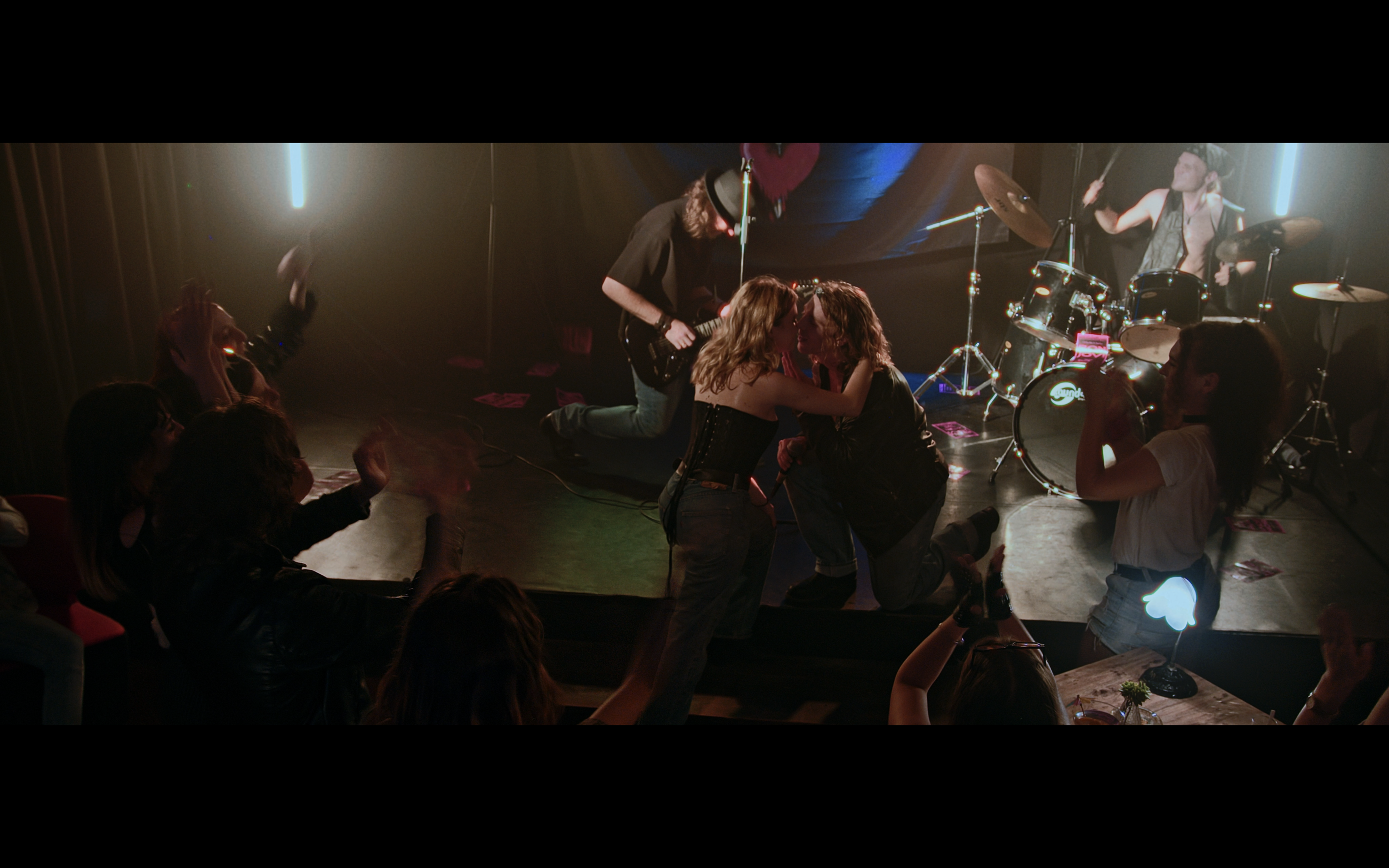 Una scena di un concerto dal vivo con persone che ballano e si divertono, mentre sul palco ci sono una band con batteria, chitarra e altri strumenti musicali.
