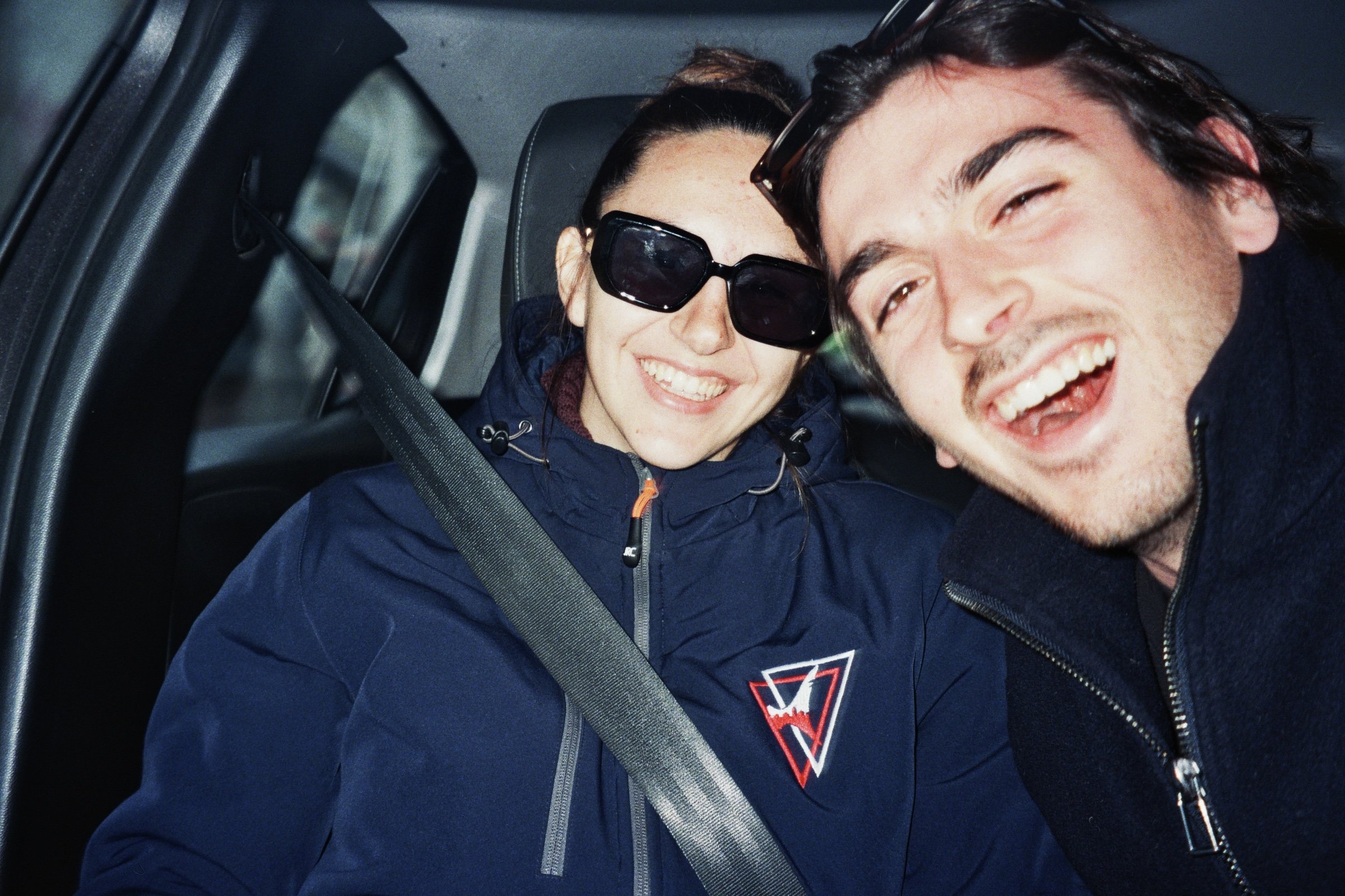 Due persone sorridenti sedute in auto, uno con occhiali da sole e vestiti sportivi, l'altro con abiti casual, scattando una foto insieme.