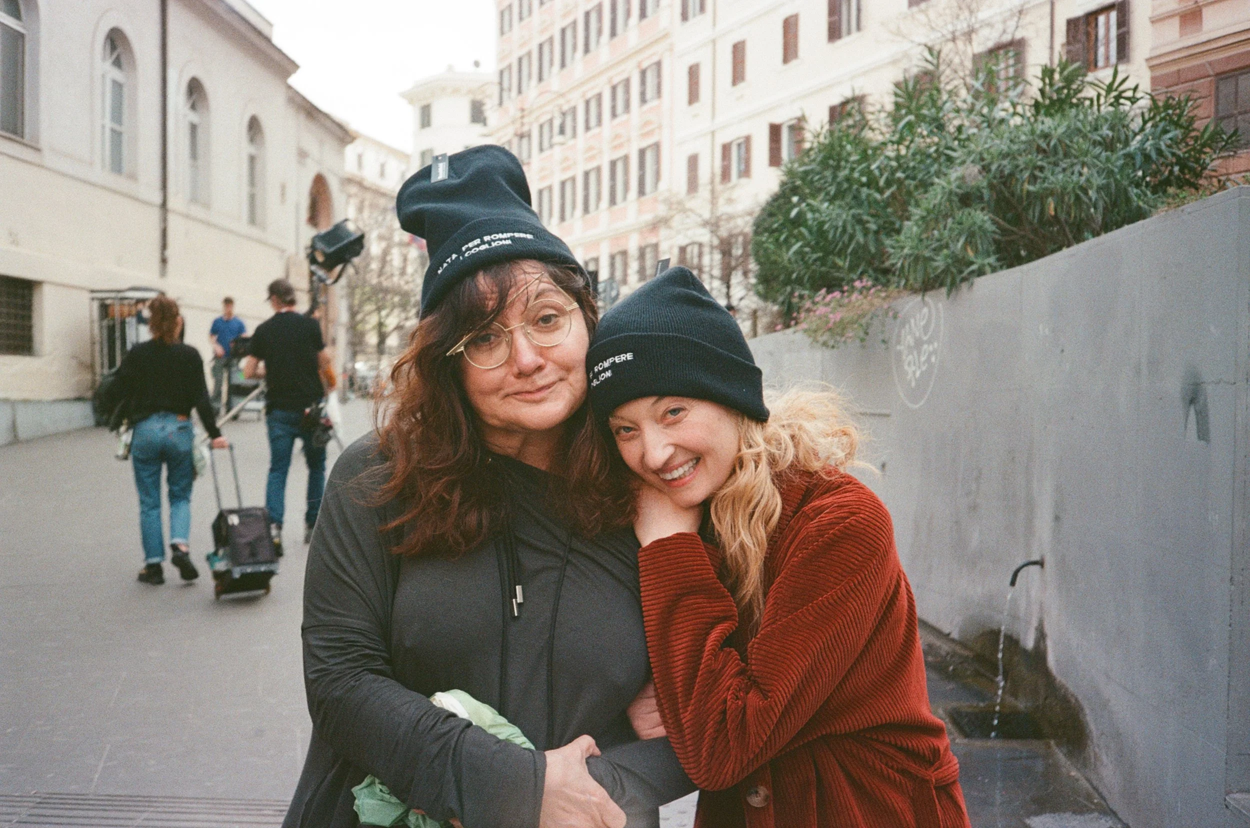 Alba Rohrwacher e Isabel Coixet sorridenti e abbracciate in strada urbana, con edifici e altre persone sullo sfondo.