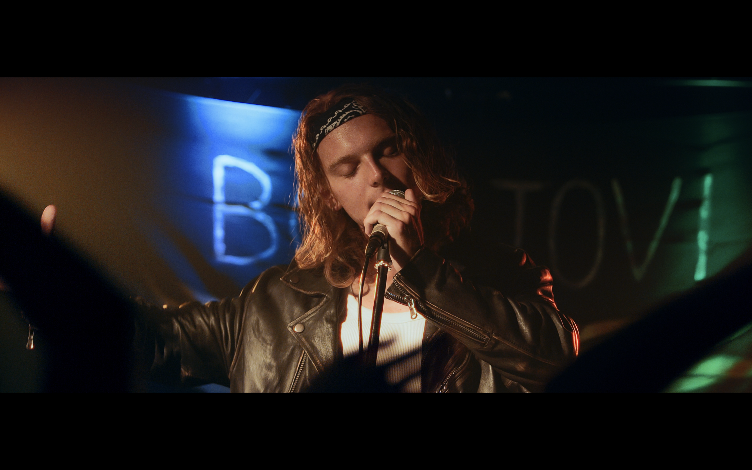 Un uomo con capelli lunghi e ricci che canta con gli occhi chiusi, indossa una giacca di pelle e una bandana nera, sullo sfondo luci colorate e un neon blu con la lettera 'B'.