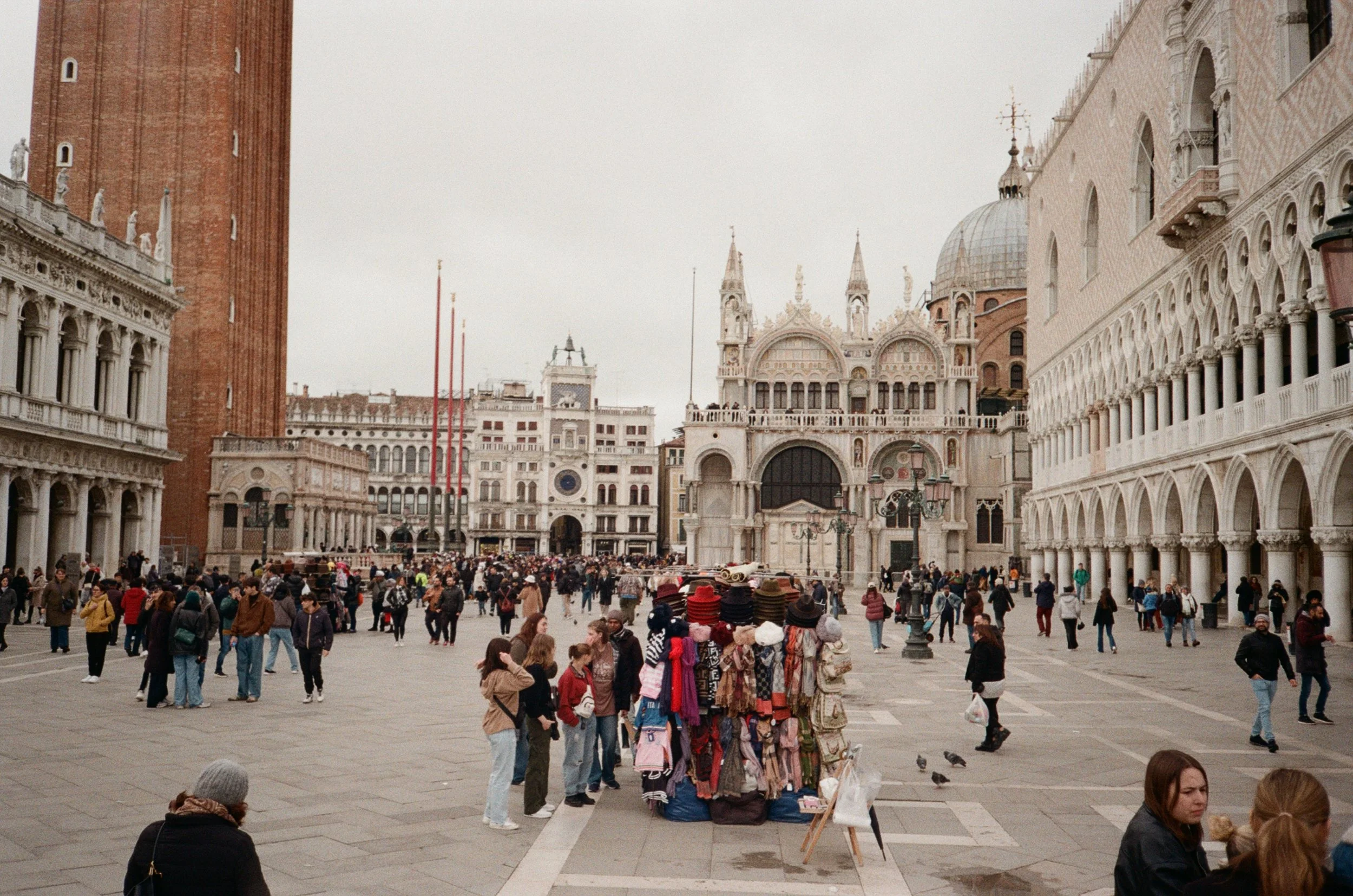 Piazza San Marco a Venezia con molte persone, un banco di vendita di cappelli colorati, e l'architettura storica con basilica e campanile.