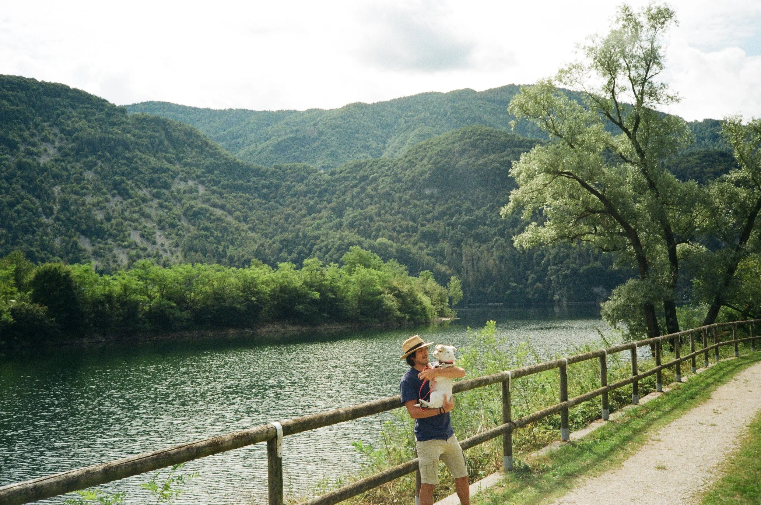 Persona che tiene un cane vicino a un lago con montagne sullo sfondo, percorso pedonale e rete di confine