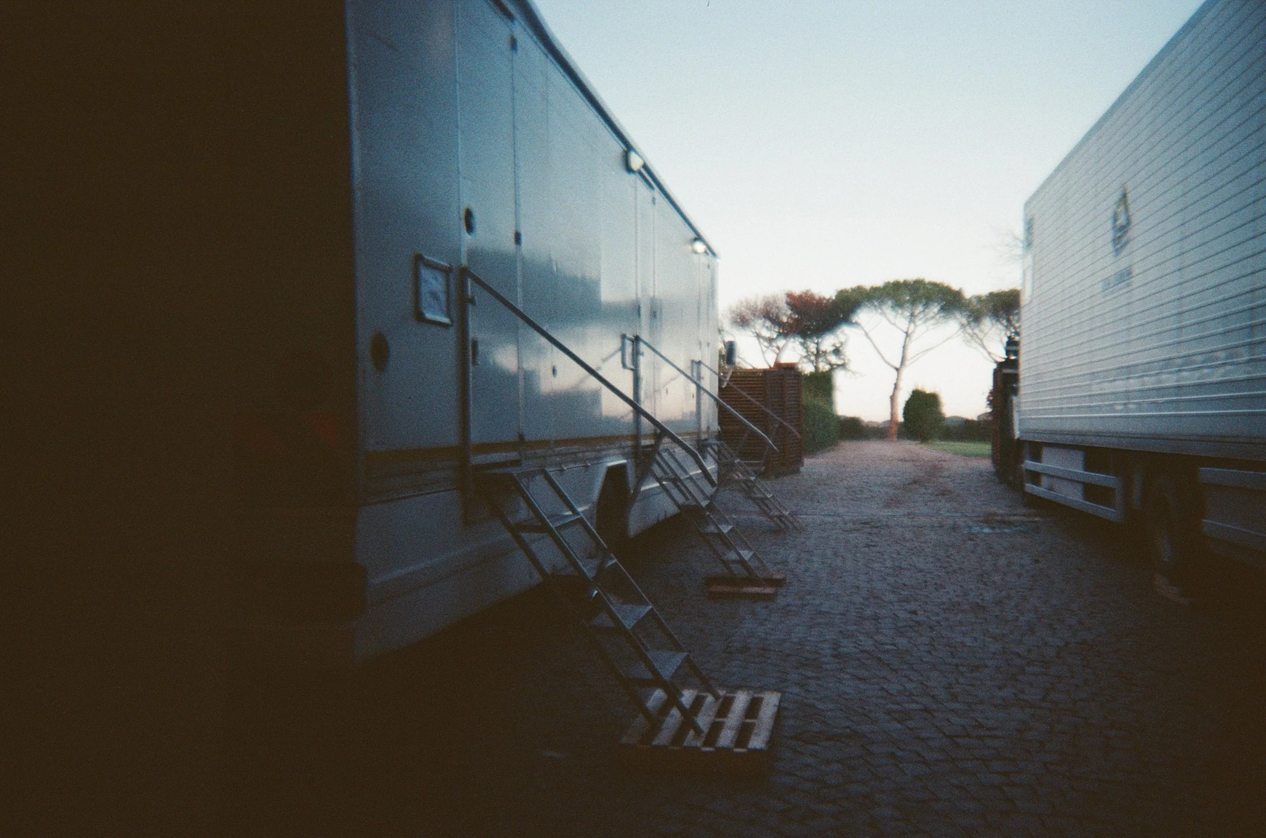 Due grandi trailer o container di camion parcheggiati su una strada di ciottoli, con alcune scale di accesso e un'area con alberi sullo sfondo.