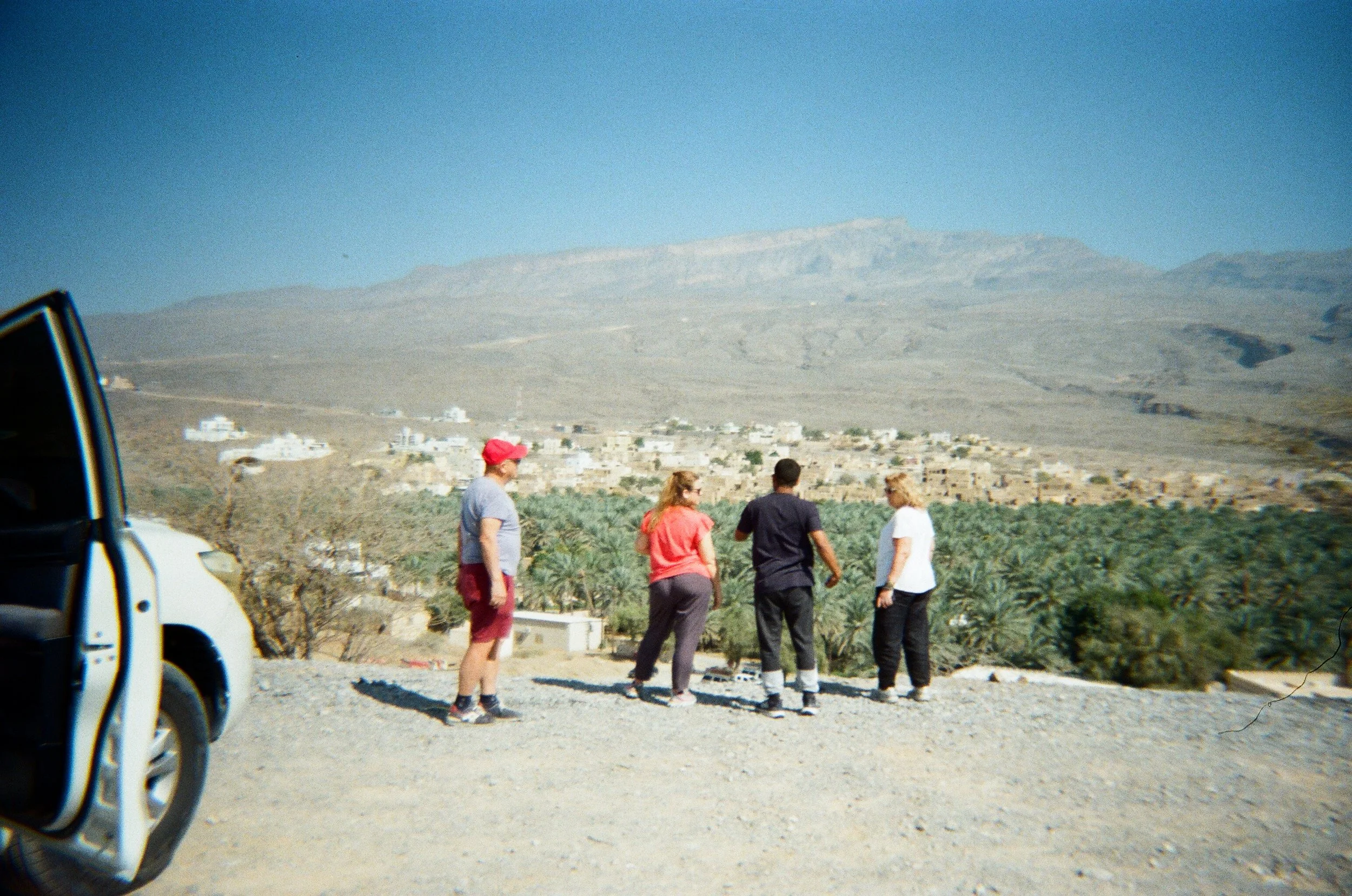 Un gruppo di quattro persone in piedi su una collina con un paesaggio desertico e un campo di palme sullo sfondo, vicino a un'auto bianca parcheggiata a sinistra.