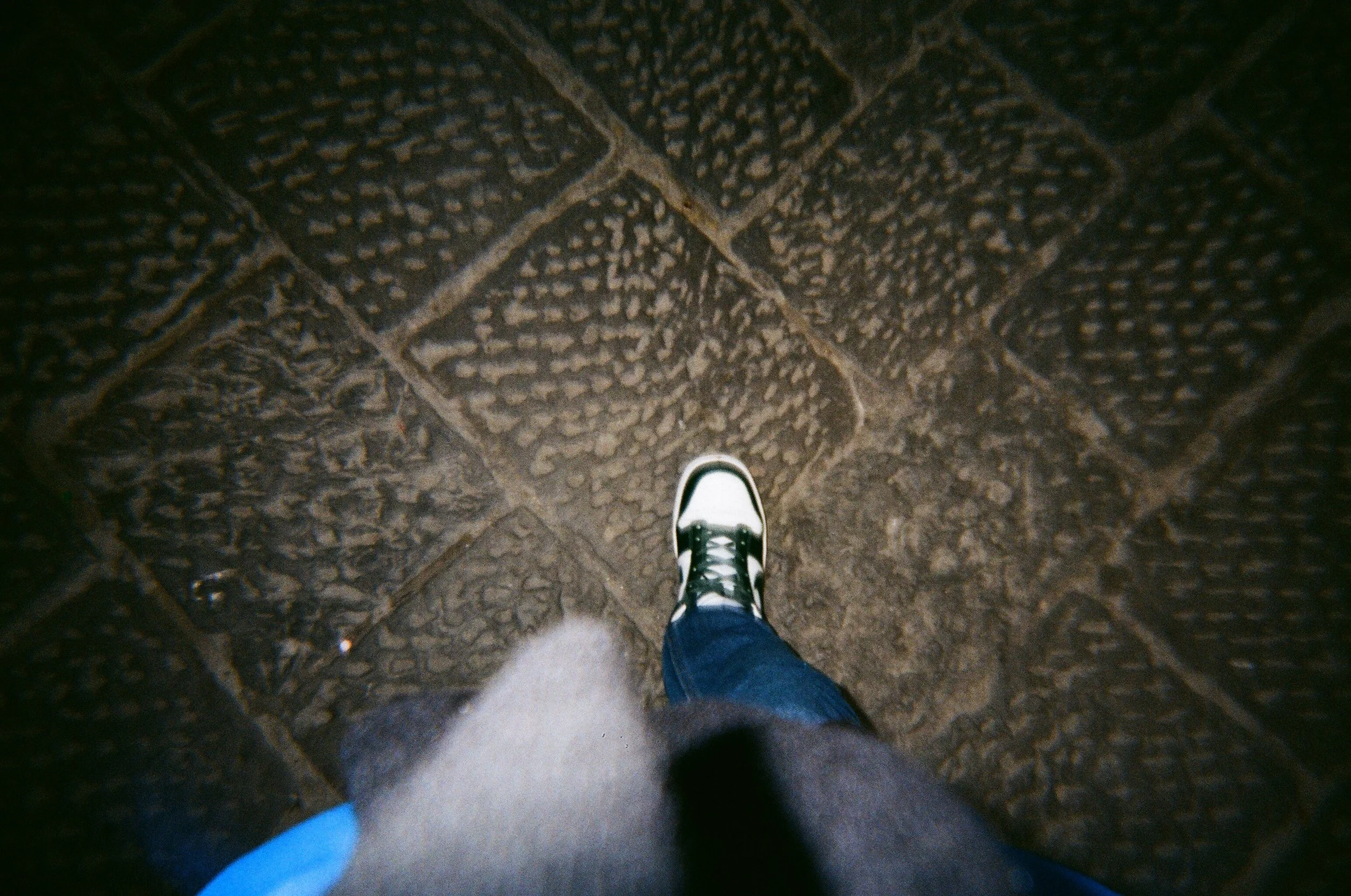 Vista dall'alto di una persona con scarpe sportive bianche e nere, pantaloni blu e cappotto grigio, che cammina sulla pavimentazione in pietra
