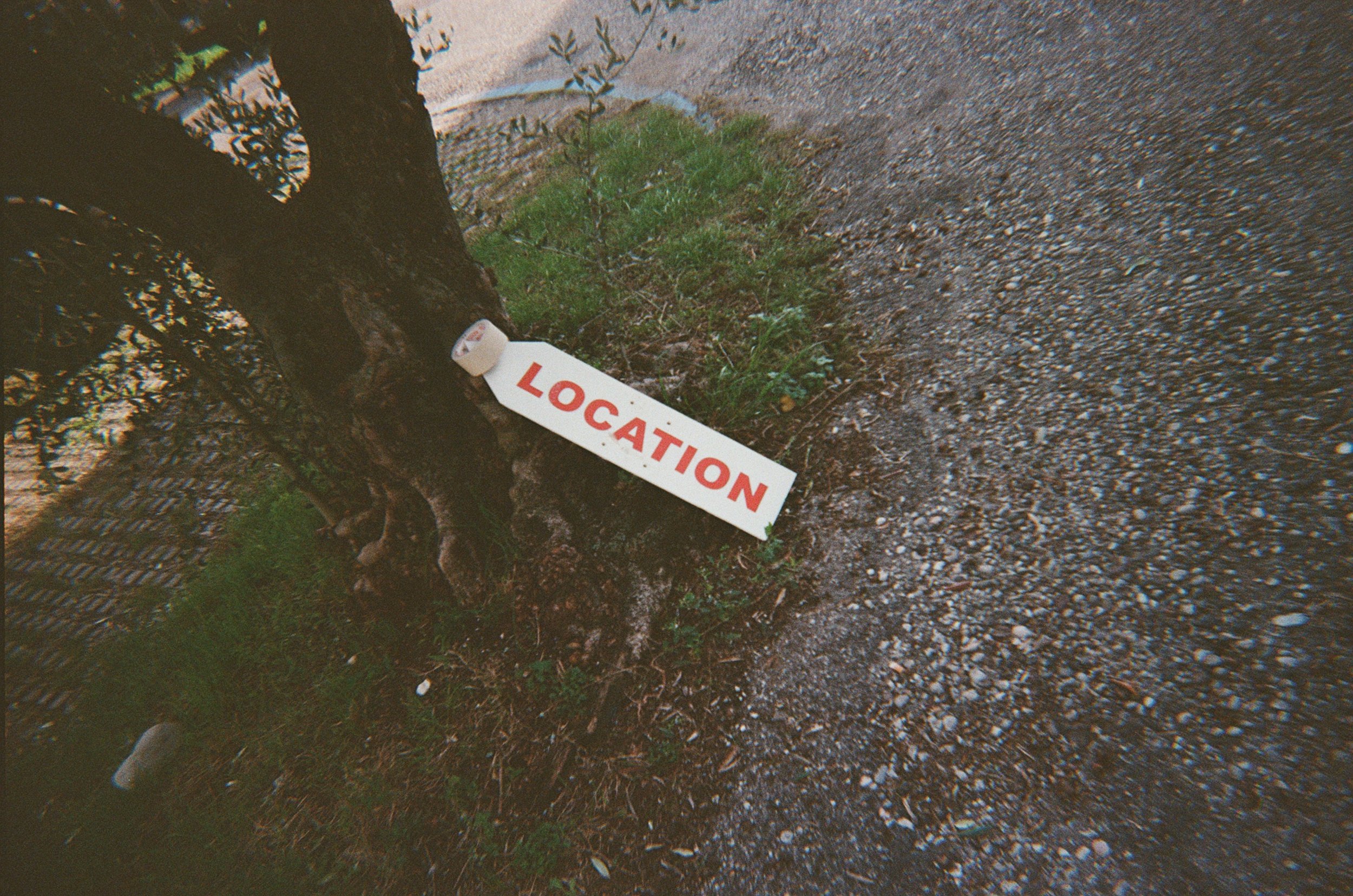 Segnale di localizzazione abbandonato a terra vicino a un albero e un marciapiede, con scritto 'LOCATION' in rosso.