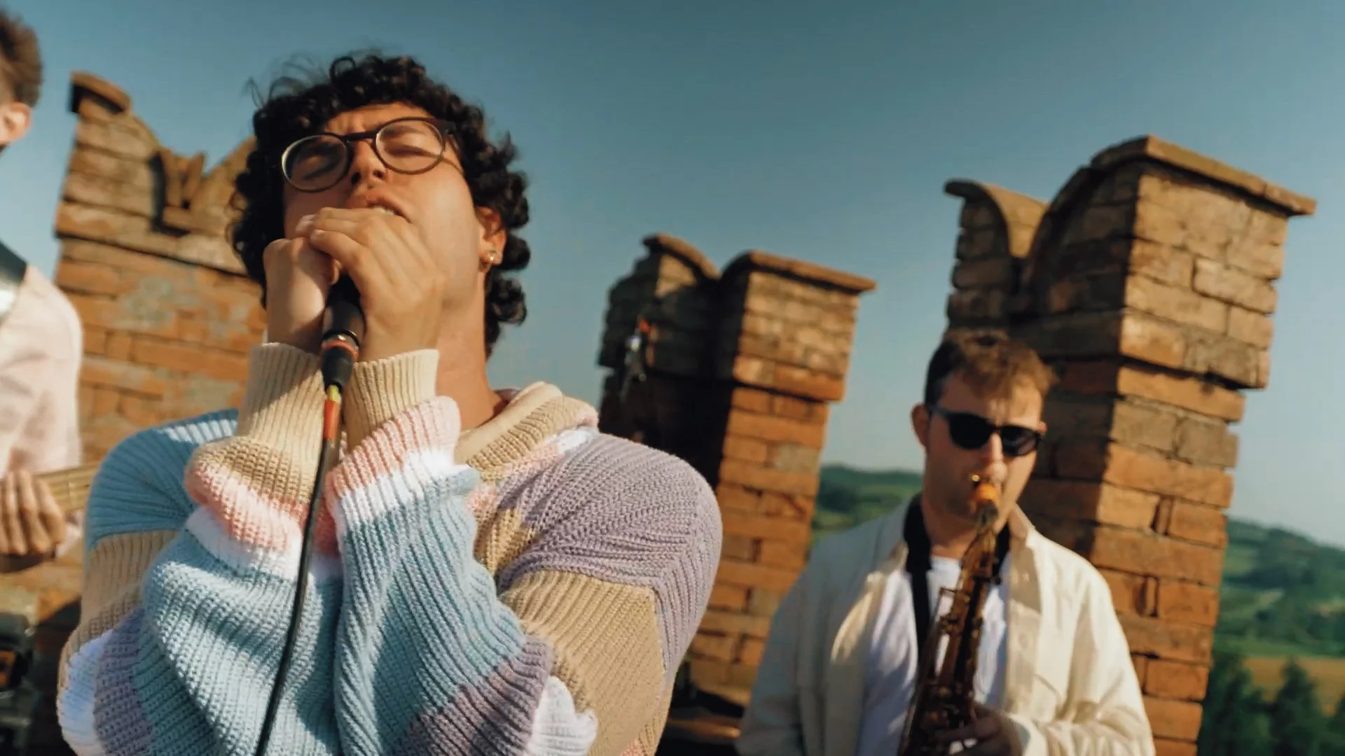 Gruppo di giovani musicisti che suonano strumenti musicali all'aperto, con sfondo di torri di mattoni e un cielo chiaro.