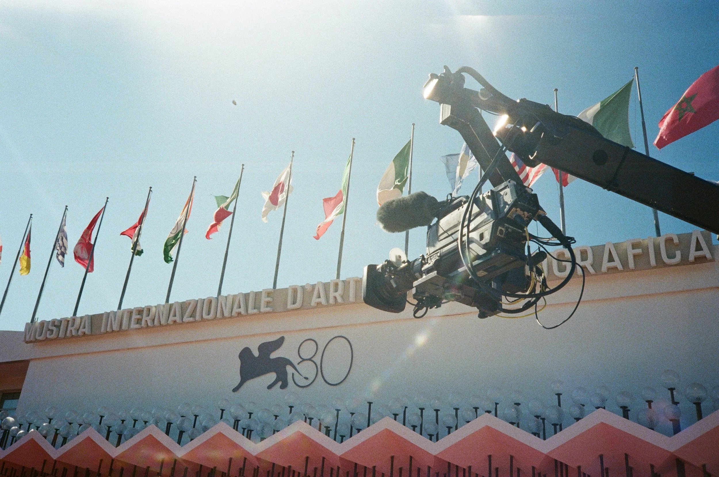 Fotografia di un'installazione all'aperto con vari drappi di bandiere di diversi paesi, sopra una struttura che recita 'Mostra Internazionale D'Arte Grafica', con un'illuminazione naturale e una macchina da presa in primo piano.