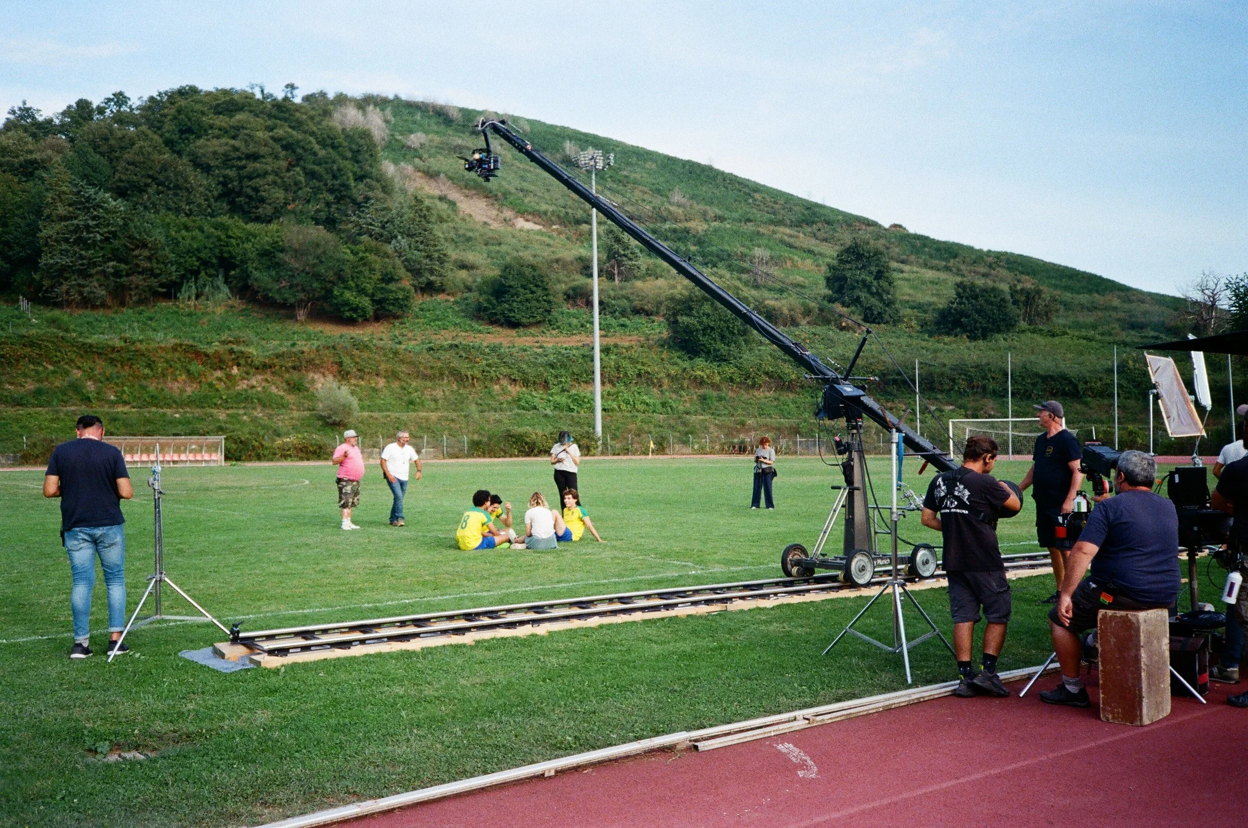 Set cinematografico in un campo sportivo con una telecamera a braccio in azione, persone sedute e in piedi che preparano o partecipano a una scena, sullo sfondo colline verdi e cielo azzurro.