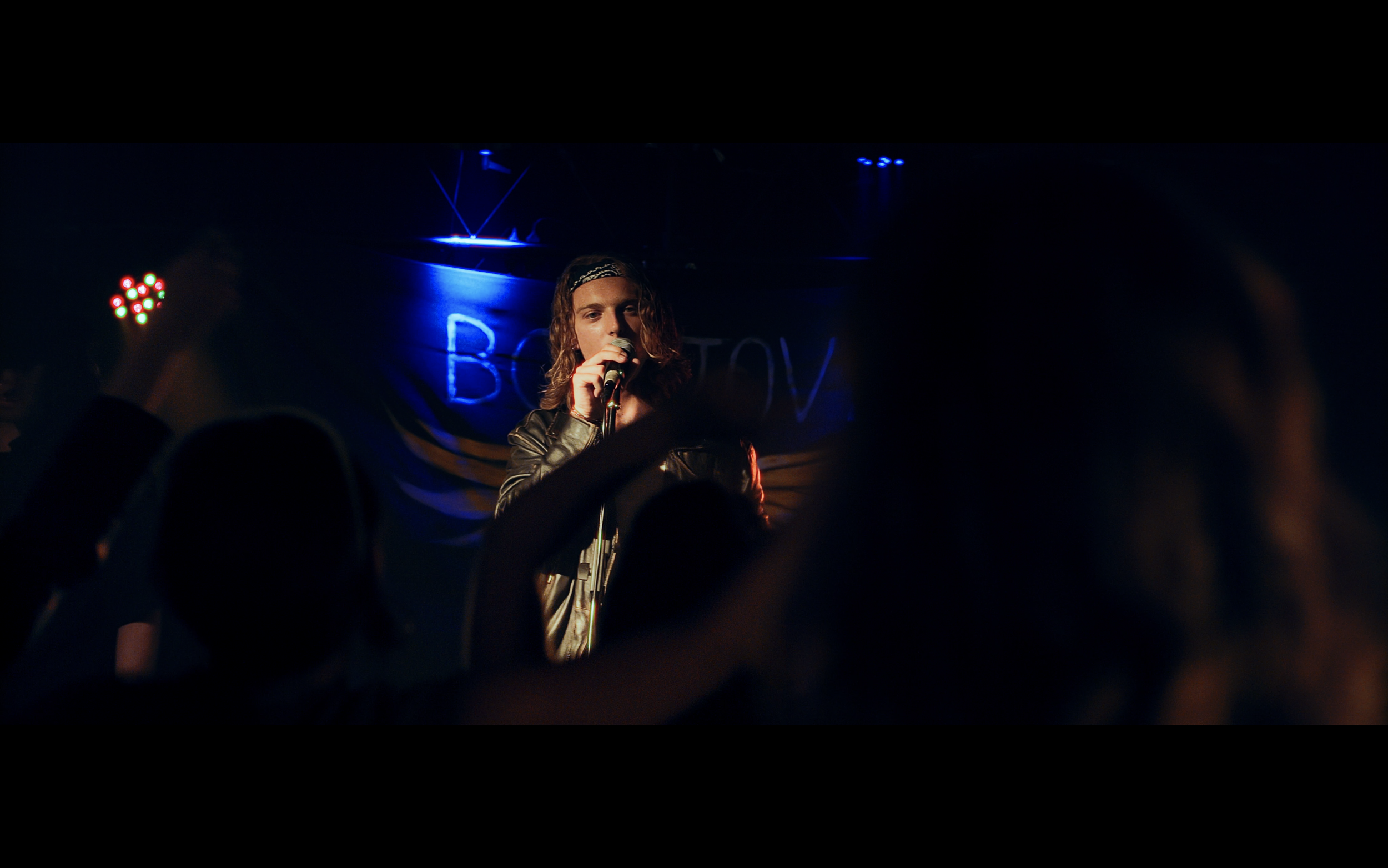 Un cantante maschile con cappello e giacca di pelle canta con un microfono di fronte a un pubblico in un locale, con luci blu e un cartellone neon in background.