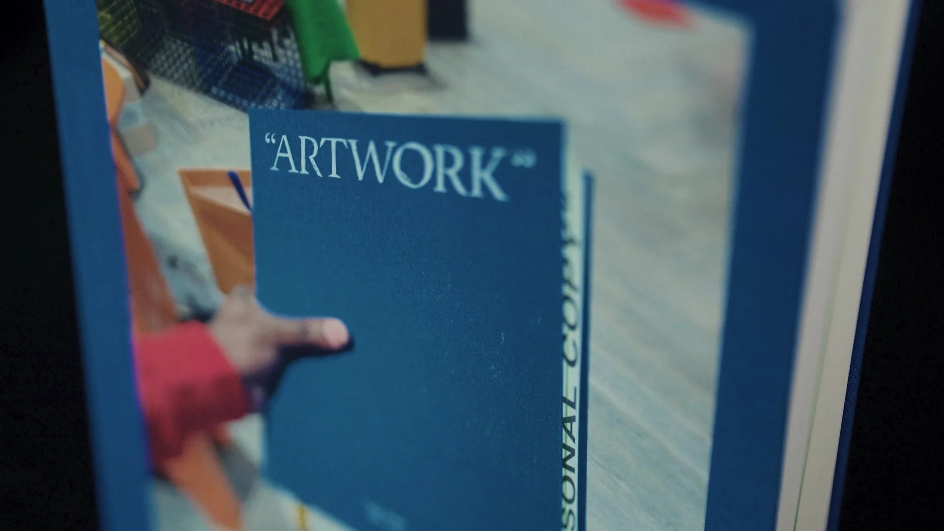 Libro con copertina blu intitolato "Artwork" con immagine di una mano e sfondo scolorito, visibile una libreria con vari libri sullo sfondo.