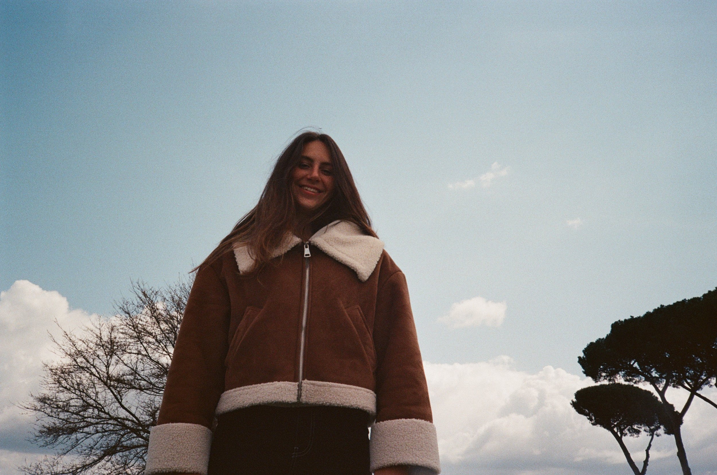 Una giovane donna sorridente indossa una giacca marrone con collo e maniche in pelliccia bianca, si trova all'aperto con cielo azzurro e nuvole, e alberi sullo sfondo.