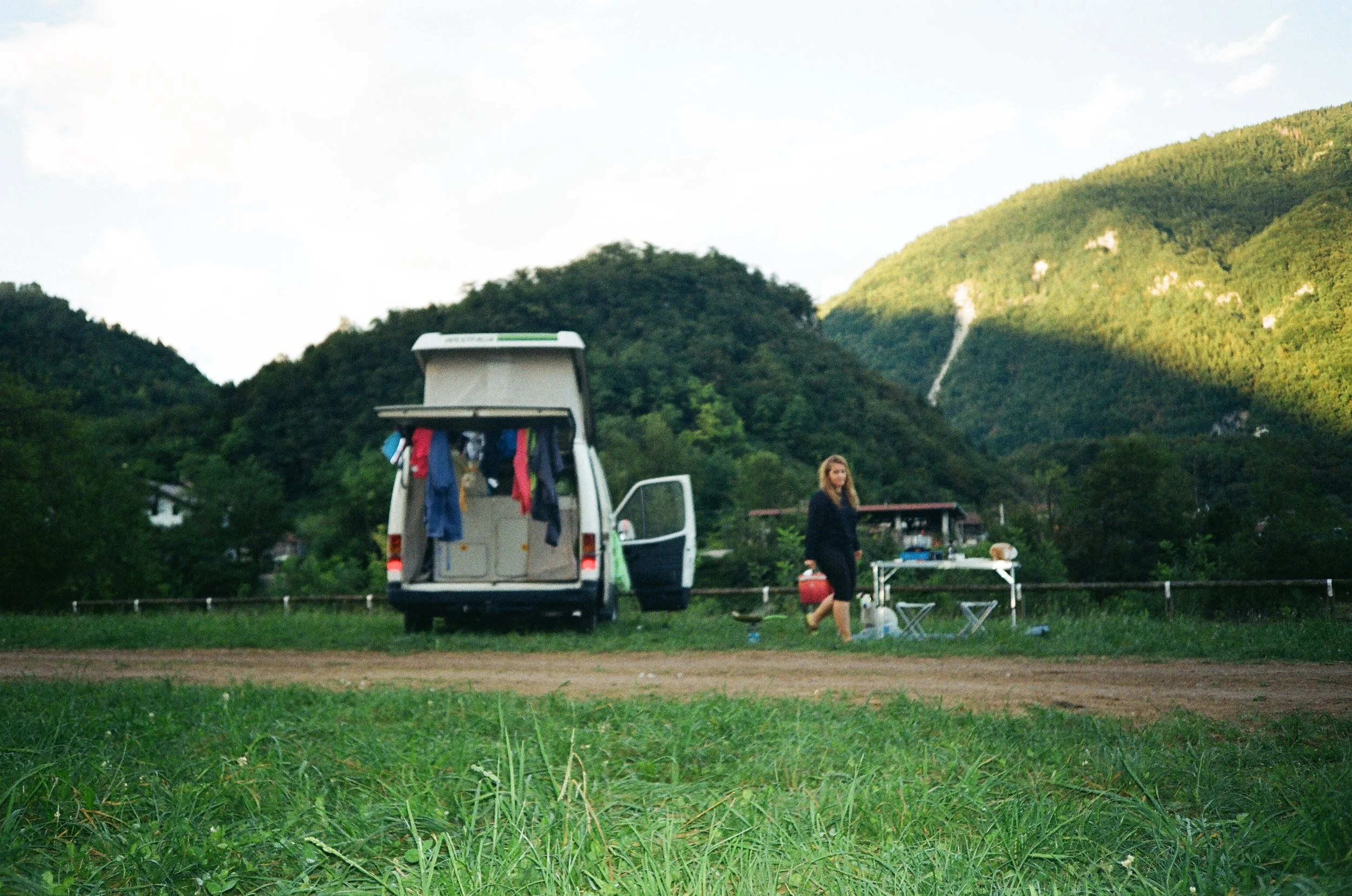 Una donna che si prepara a cucinare davanti a un parcheggio di camper in una campagna verde con montagne sullo sfondo.