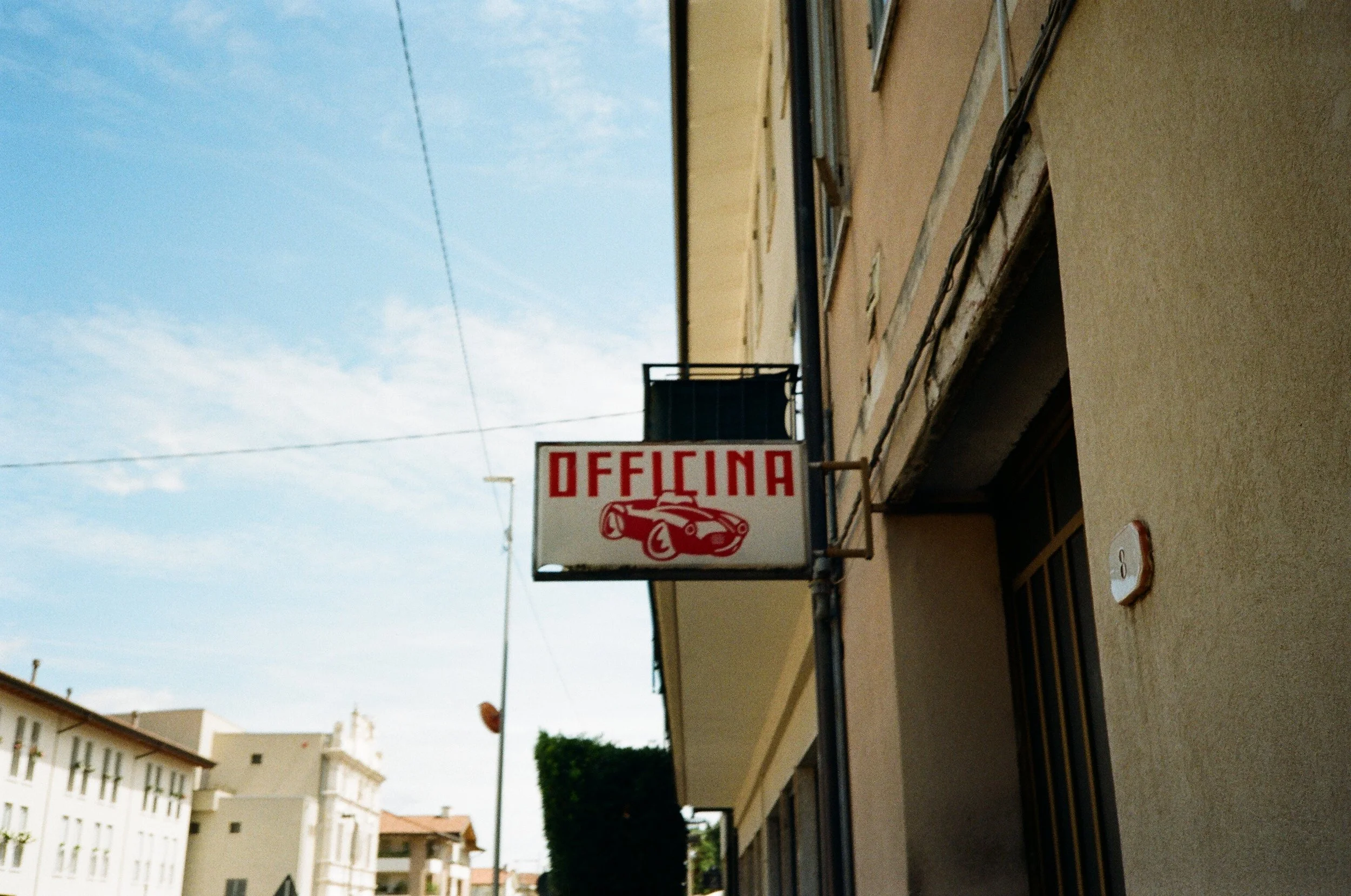 Cartello con scritta 'Officina' e disegno di un'auto sportiva rossa davanti a un edificio a più piani in una strada urbana.