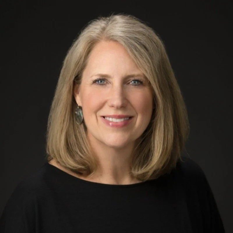 Headshot of a woman with shoulder-length blonde hair, wearing a black top and earrings, smiling against a dark background.
