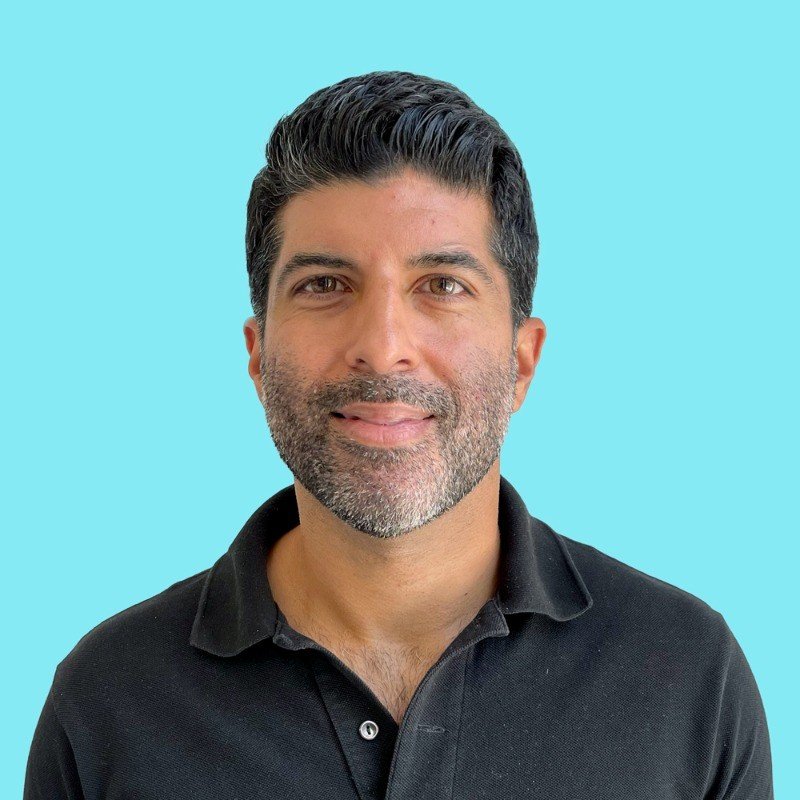 A man with dark hair, gray beard, wearing a black polo shirt, smiling against a light blue background.
