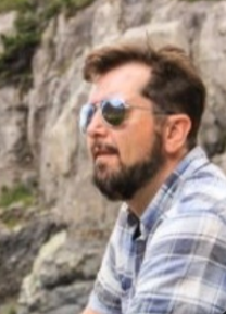 Chris Anthony is shown with a beard and sunglasses, sitting outdoors near rocks, wearing a plaid shirt.