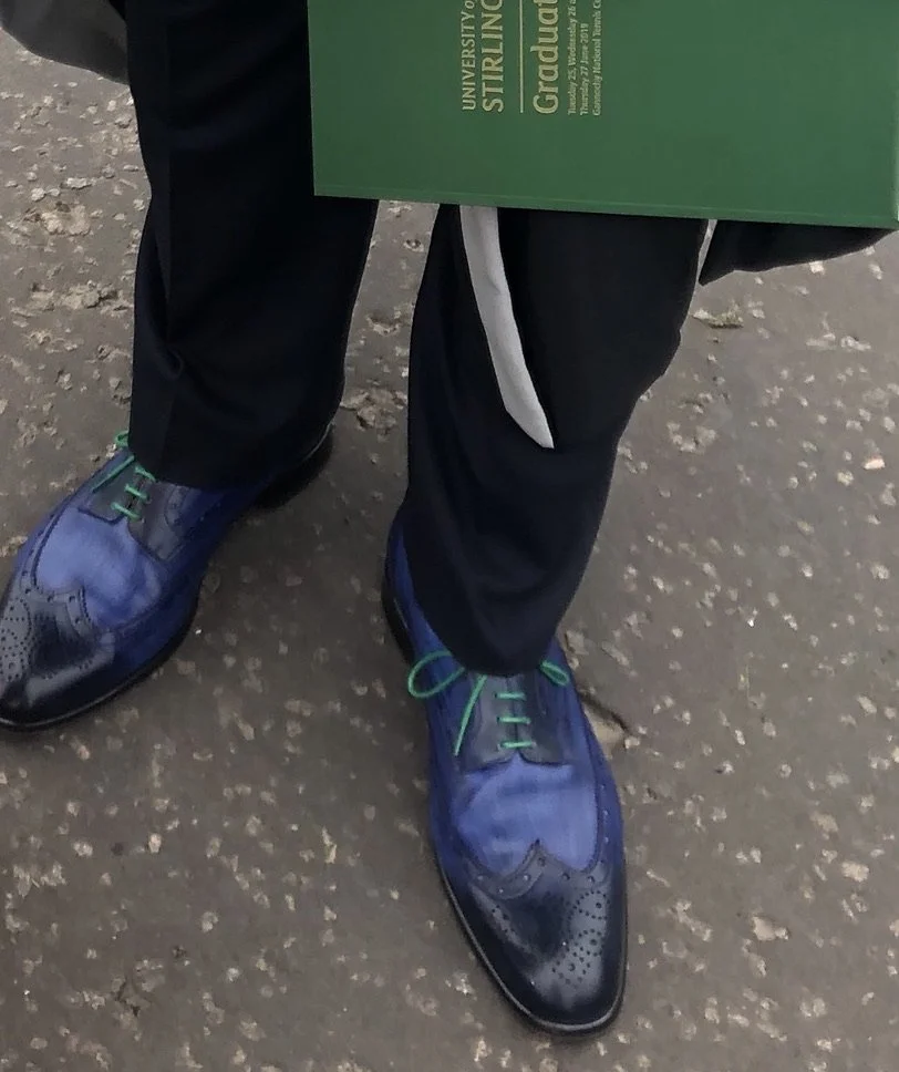 Close-up of a person wearing black pants, purple and black dress shoes with green laces, and holding a green graduation certificate from the University of Stirling.