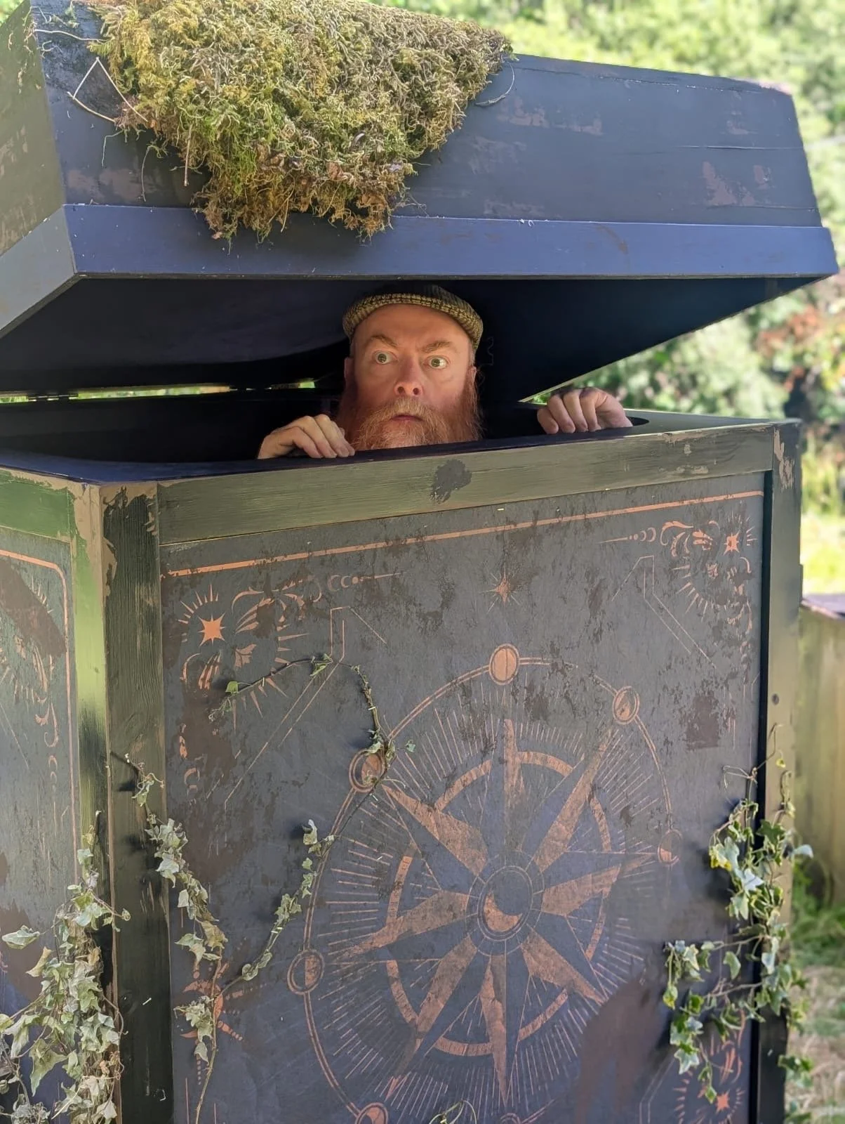 A man with a beard and a hat peeking out from a decorated outdoor box or cabinet, with a large black cover or lid above him and greenery in the background.