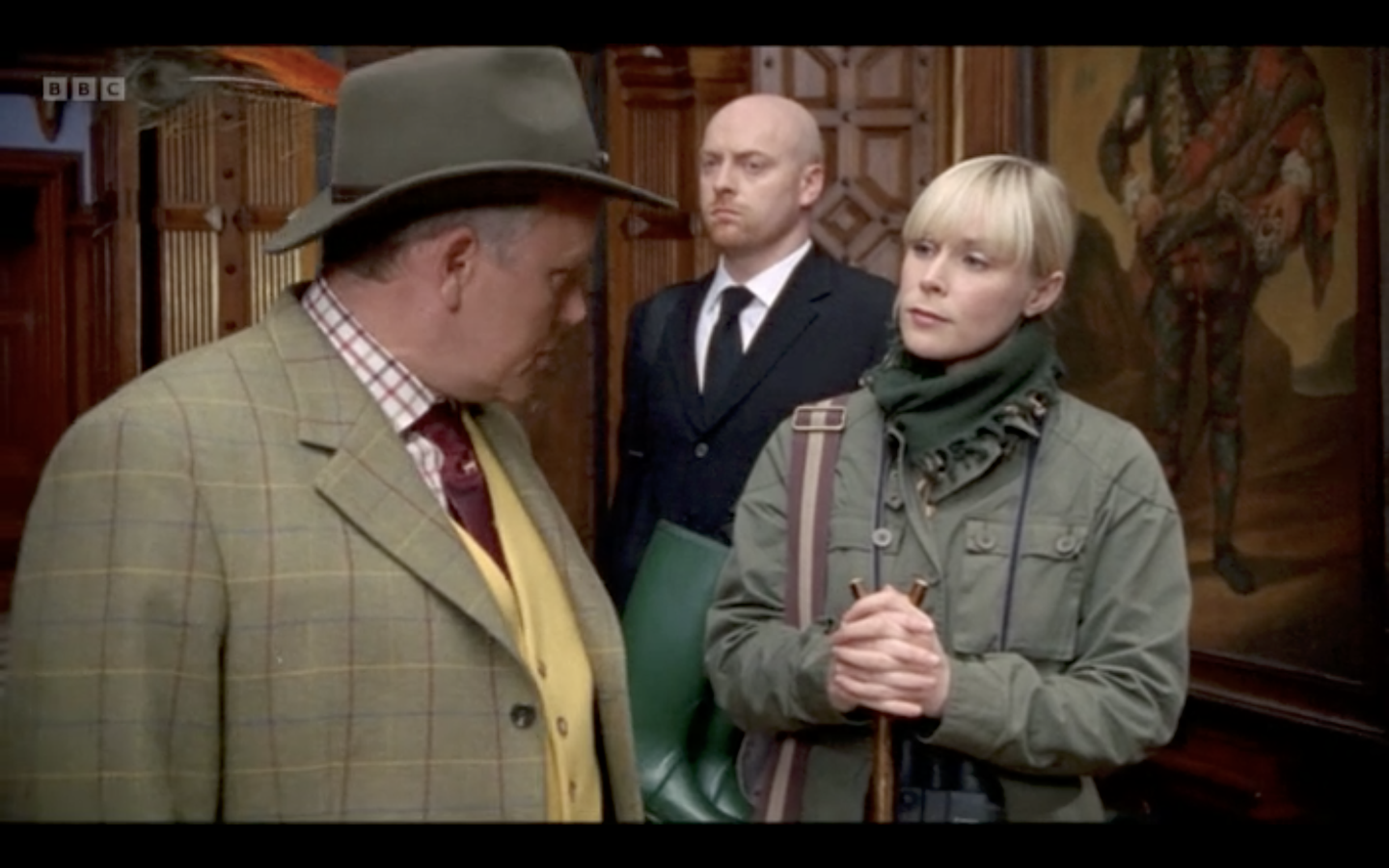 Three people are indoors, engaged in a conversation. The person on the left is wearing a light-colored blazer with a checkered shirt and a large hat. The person on the right is a young woman with short blonde hair, wearing a green jacket and holding a small object. The person in the middle is a bald man with a serious expression, dressed in a dark suit and white shirt, standing between the other two.