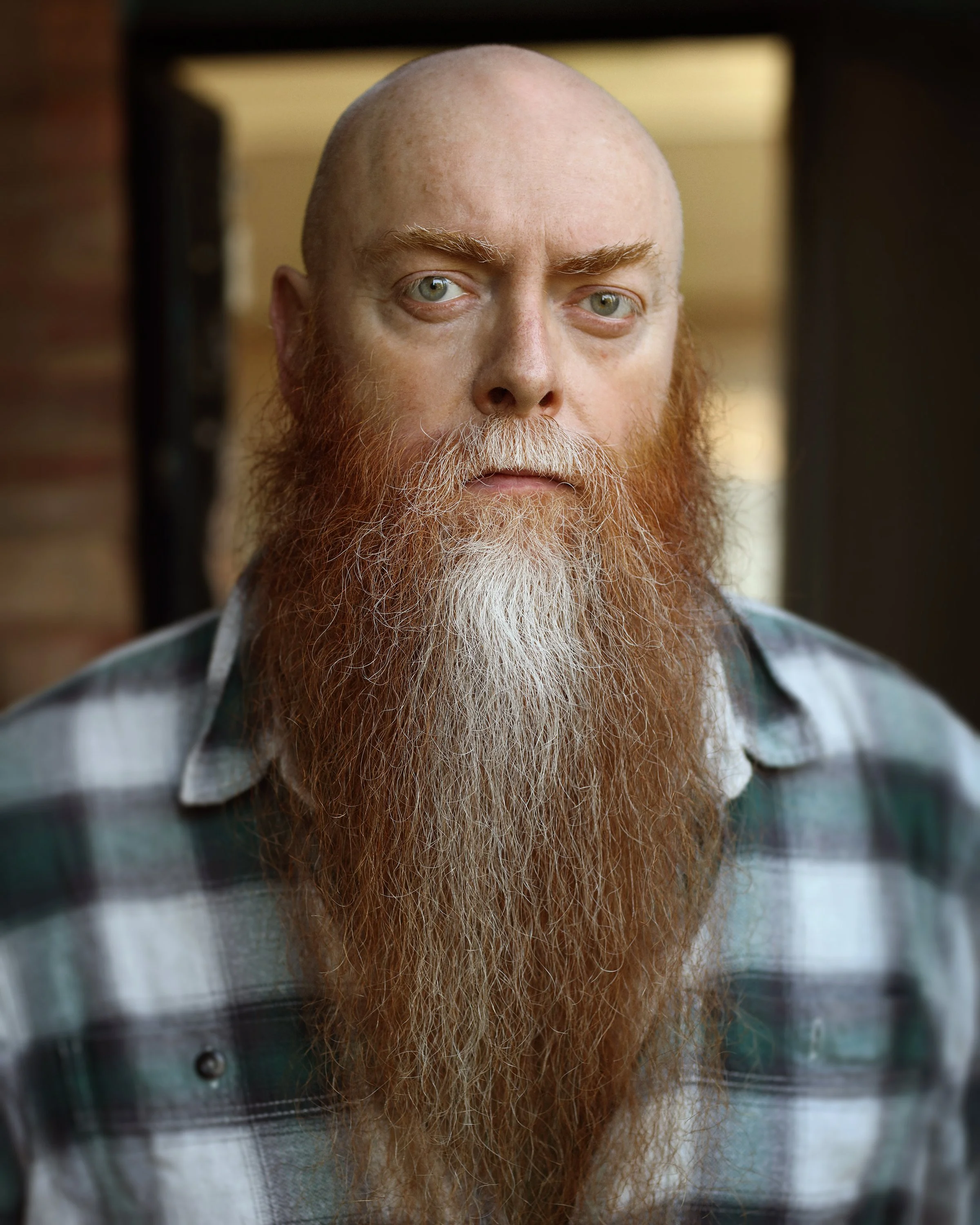 Close-up of a man with a bald head, blue eyes, and a long red beard wearing a flannel shirt, looking directly at the camera.