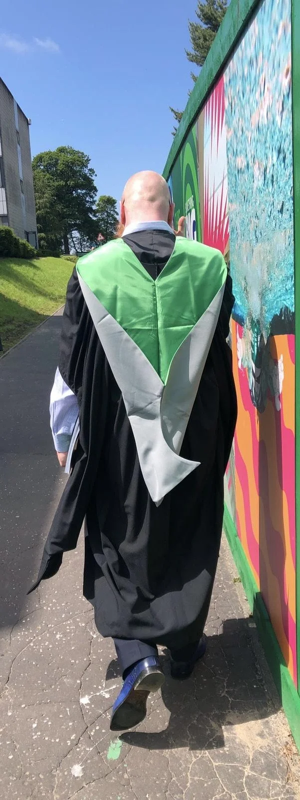 A person dressed in graduation attire walking outdoors on a sunny day with a colorful mural to the right and greenery in the background.