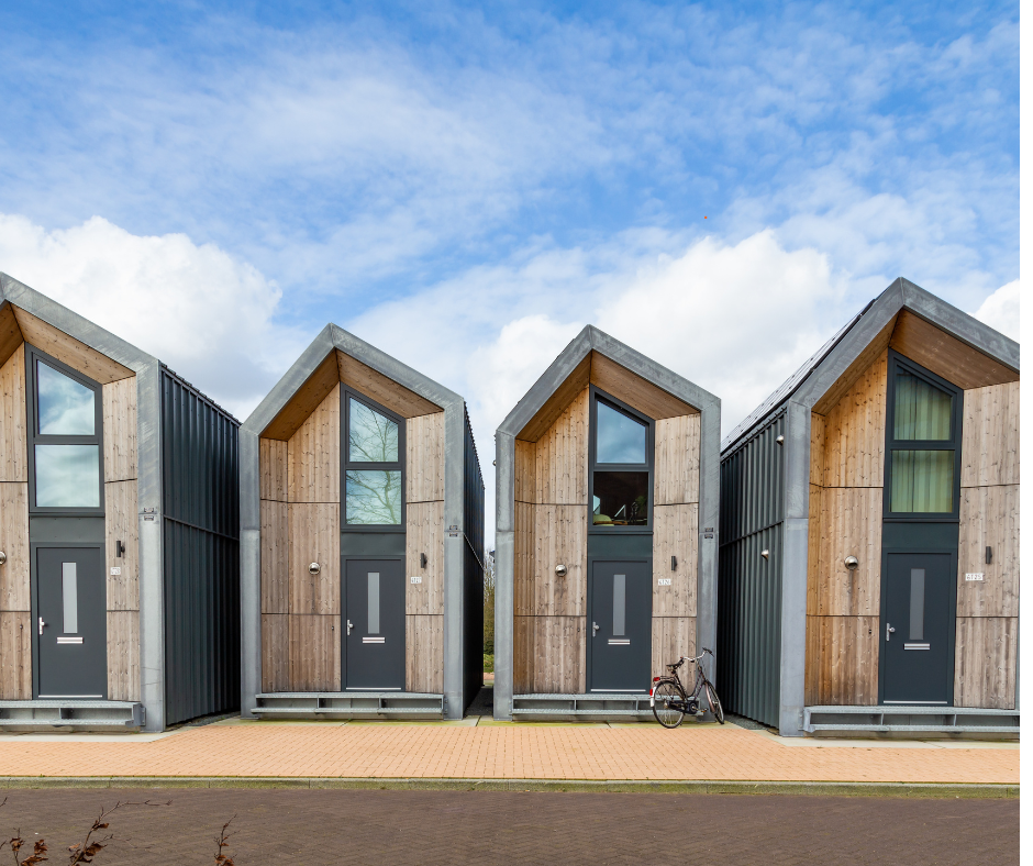 Three modern houses with wooden and metal facades, each with a gray door, large windows, and pitched roofs, located in a suburban area with a brick sidewalk and a bicycle parked outside.