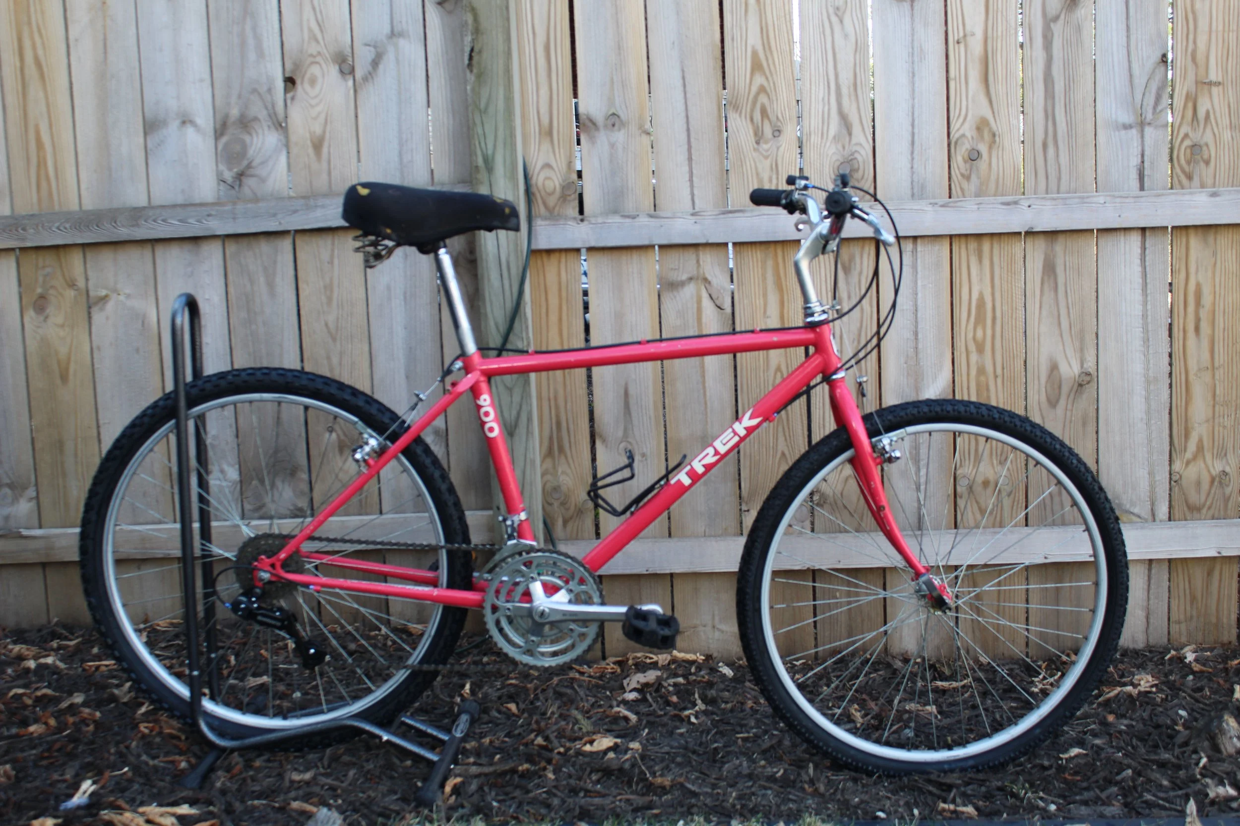 Red Trek mountain bike with black seat, in front of wooden fence, secured to a bike rack.