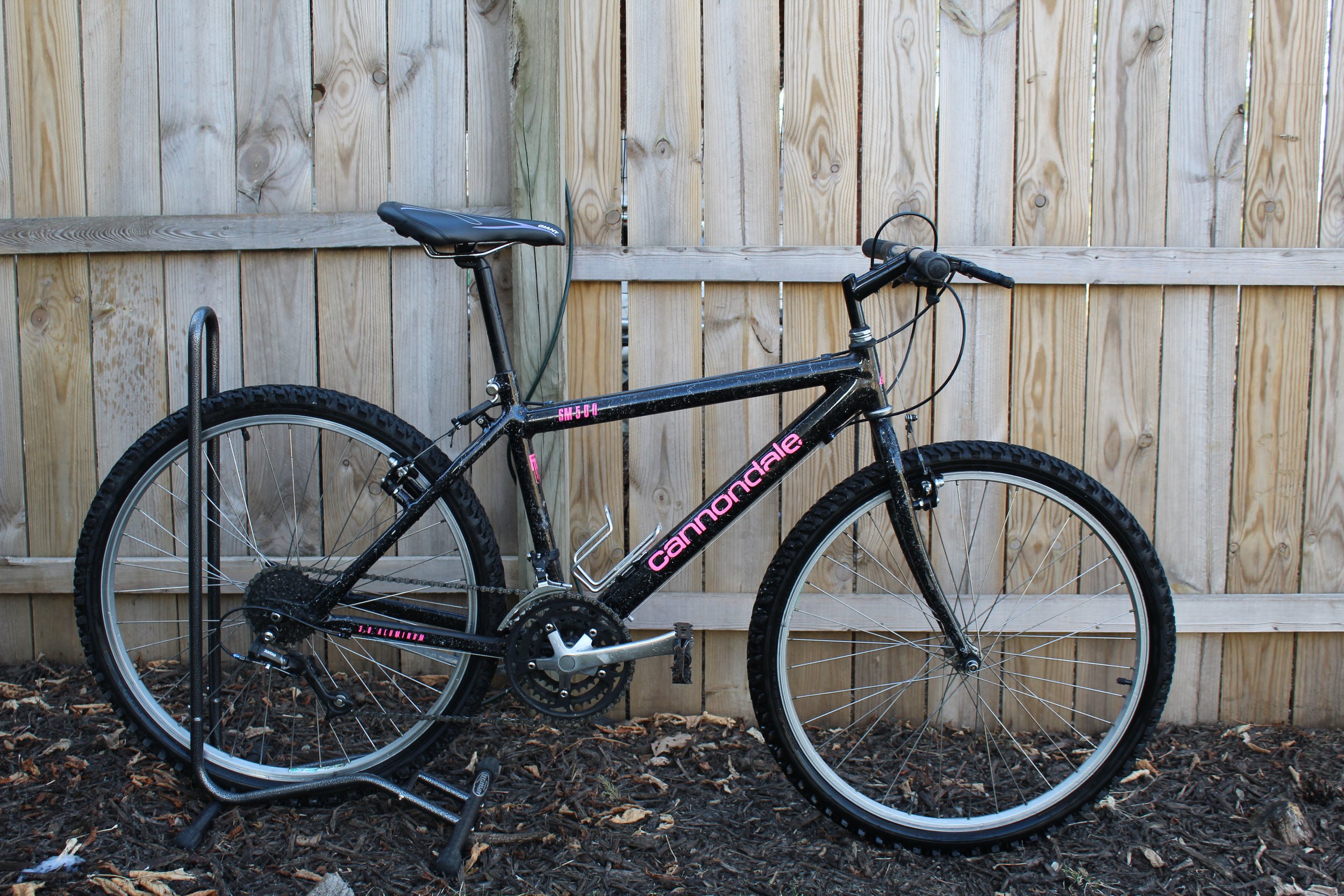 Black mountain bike with pink lettering, parked on a bike rack, wooden fence background.