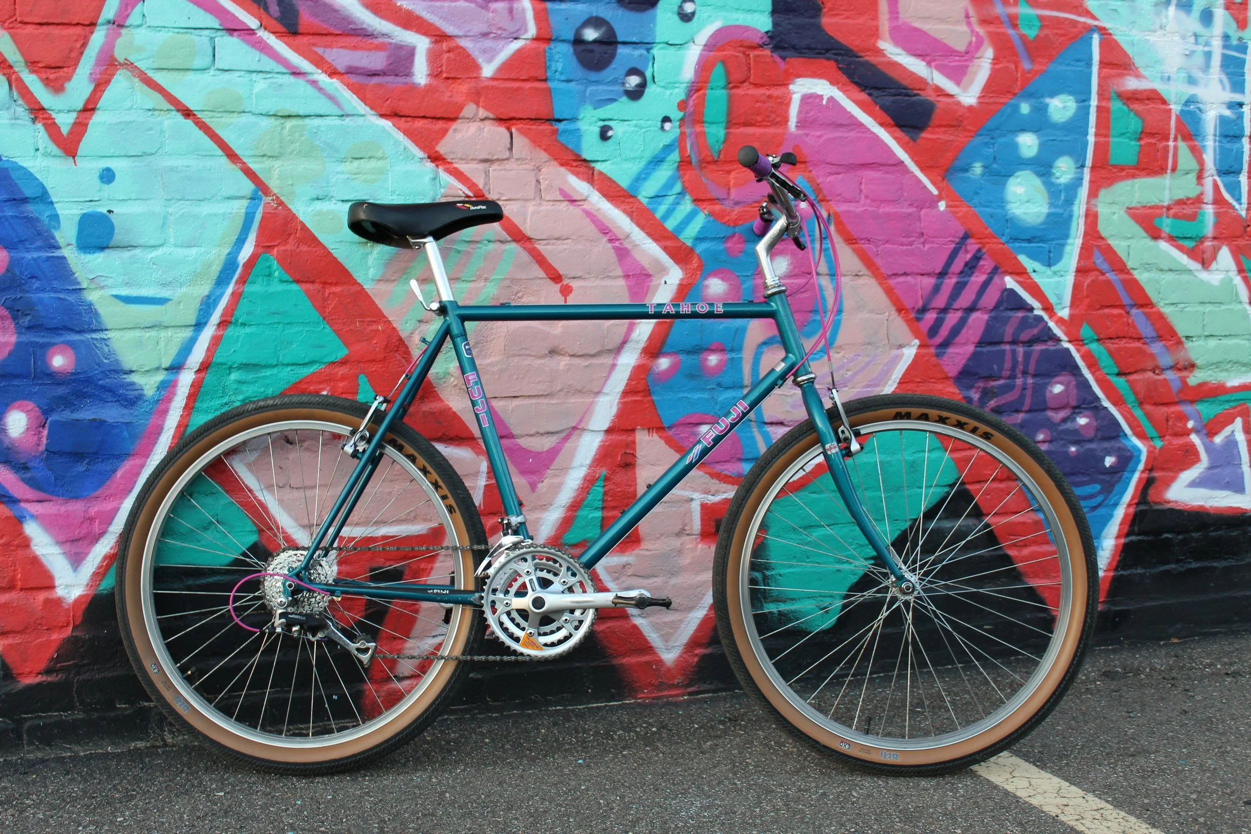 Vintage bicycle with teal frame and tan tires leaning against a colorful graffiti brick wall.