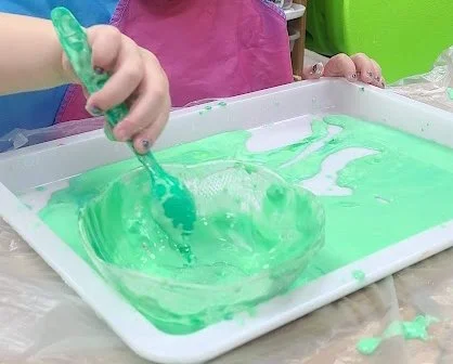 A child mixing colorful oobleck with their hands in a tray, showing both liquid and solid textures.