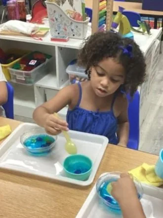 A young child sits at a table using a pipette to transfer blue water between two small glass bowls on a tray, surrounded by other bowls of primary colors.
