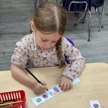 Preschool child cutting and assembling strips of printed fossil images to create a colorful, wearable fossil bracelet during a hands-on STEAM activity.