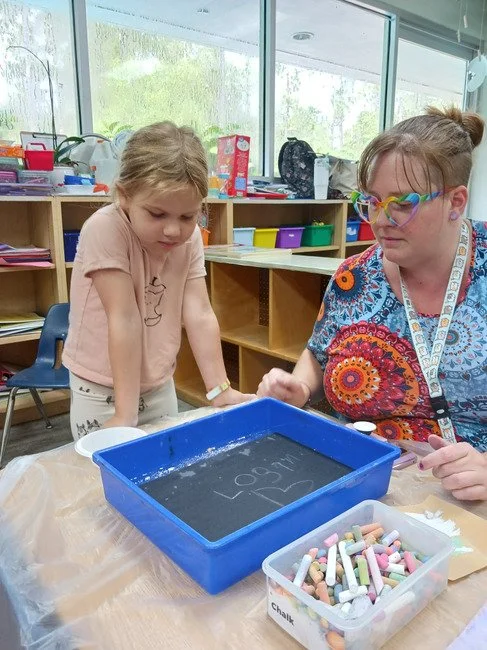 A young girl and a woman, possibly a teacher, are sitting at a table in a classroom with large windows. The girl is leaning over a blue tray filled with water, and the woman is holding chalk while looking at the water. There is a container of chalk o