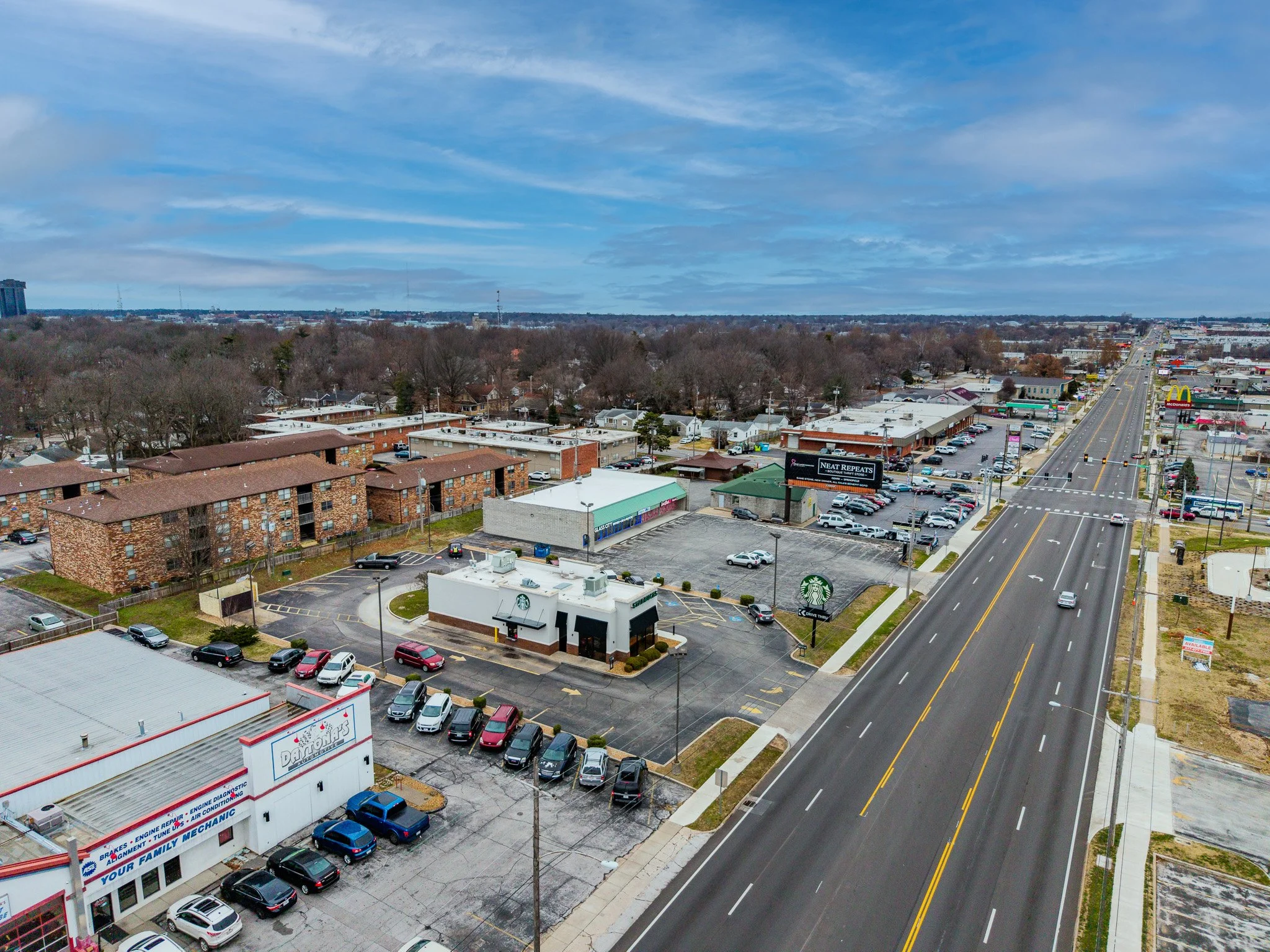 An aerial view of a commercial area with parking lots, restaurants, and stores along a wide road under a partly cloudy sky.