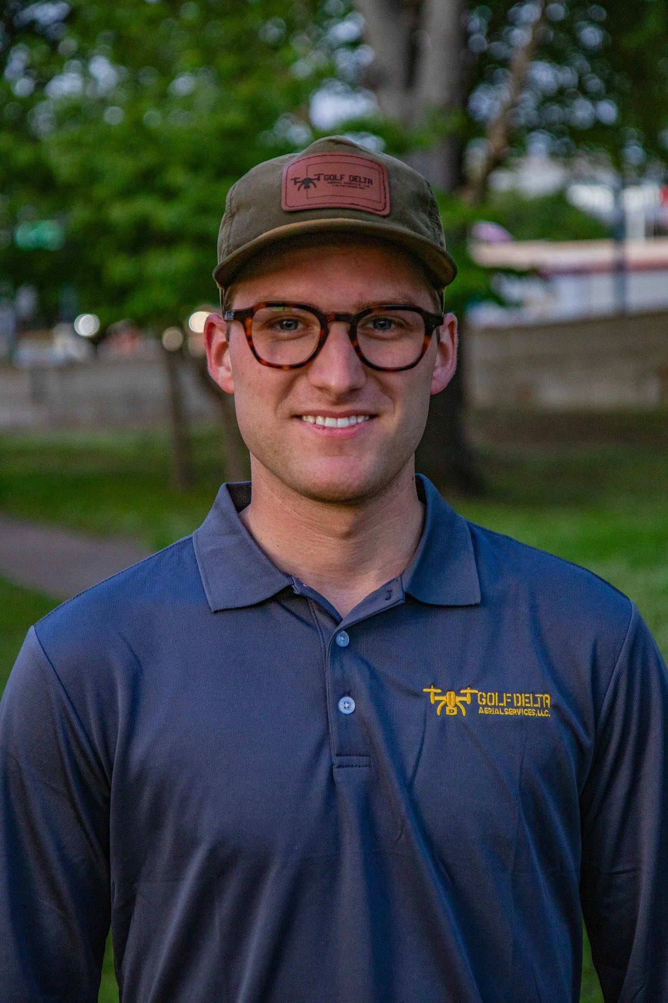 A young man with glasses and a cap, smiling outdoors in a park, wearing a blue polo shirt with 'Golf Delta' embroidered on it.