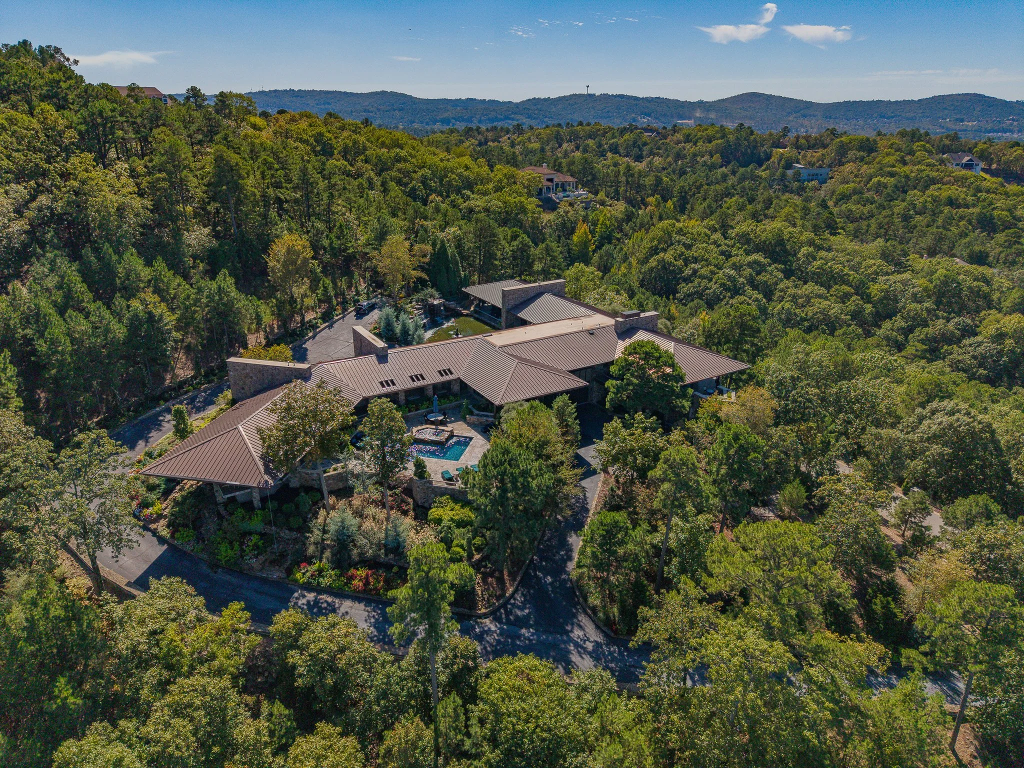 Aerial view of a large house nestled among trees in a hilly landscape, with a swimming pool and surrounding deck in the backyard.