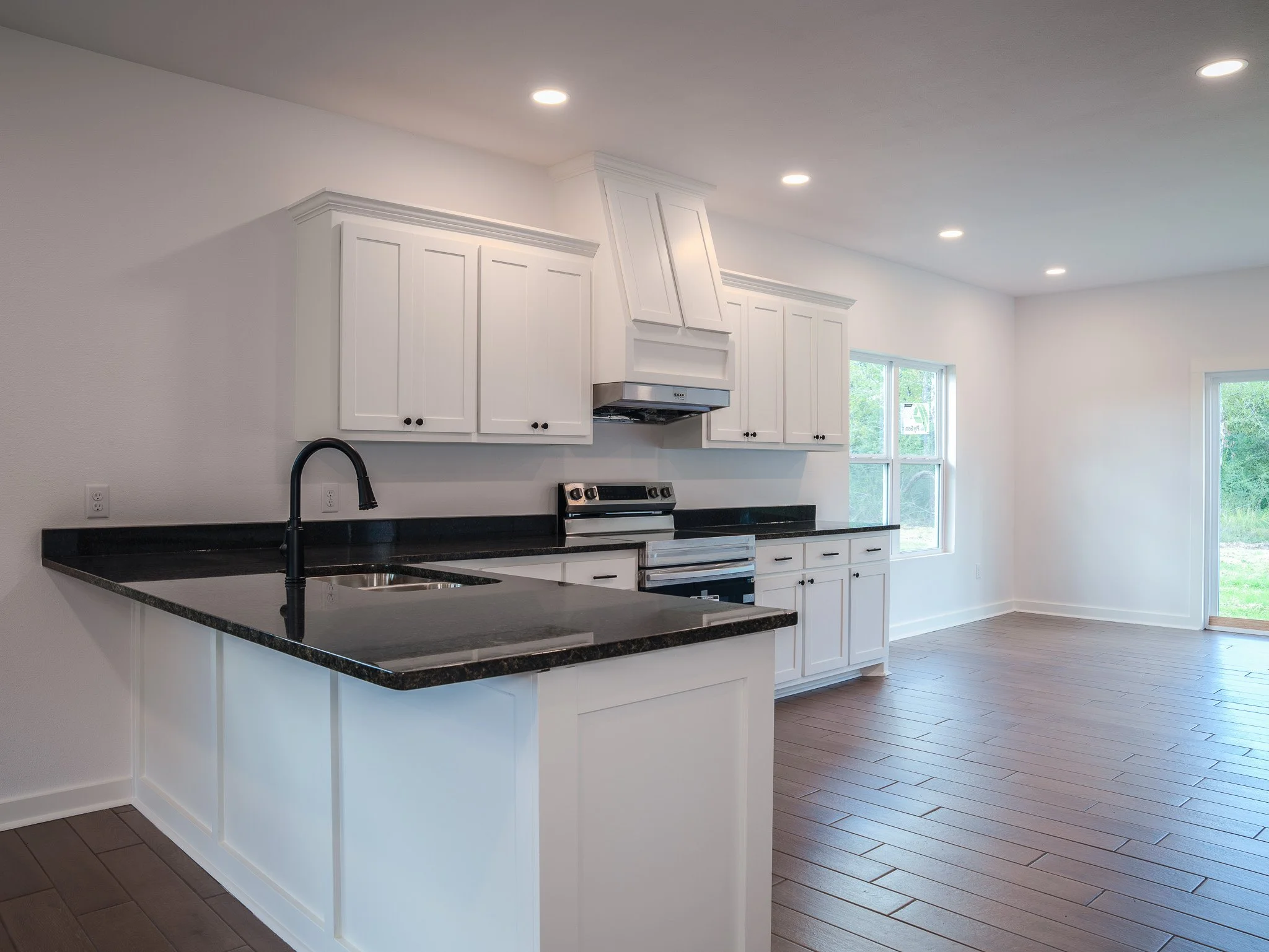 Modern kitchen with white cabinets, black countertops, stainless steel stove, black faucet, and large windows with a view of green outdoor space.