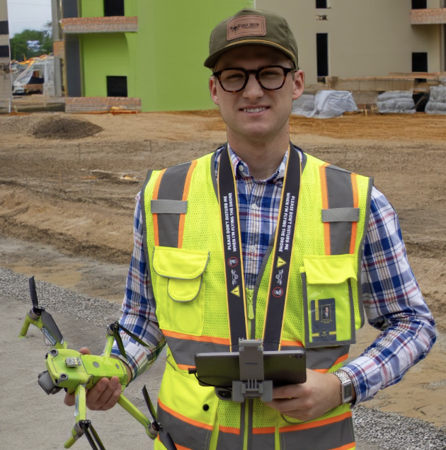 A man in safety vest and glasses standing at a construction site holding a drone and a tablet.