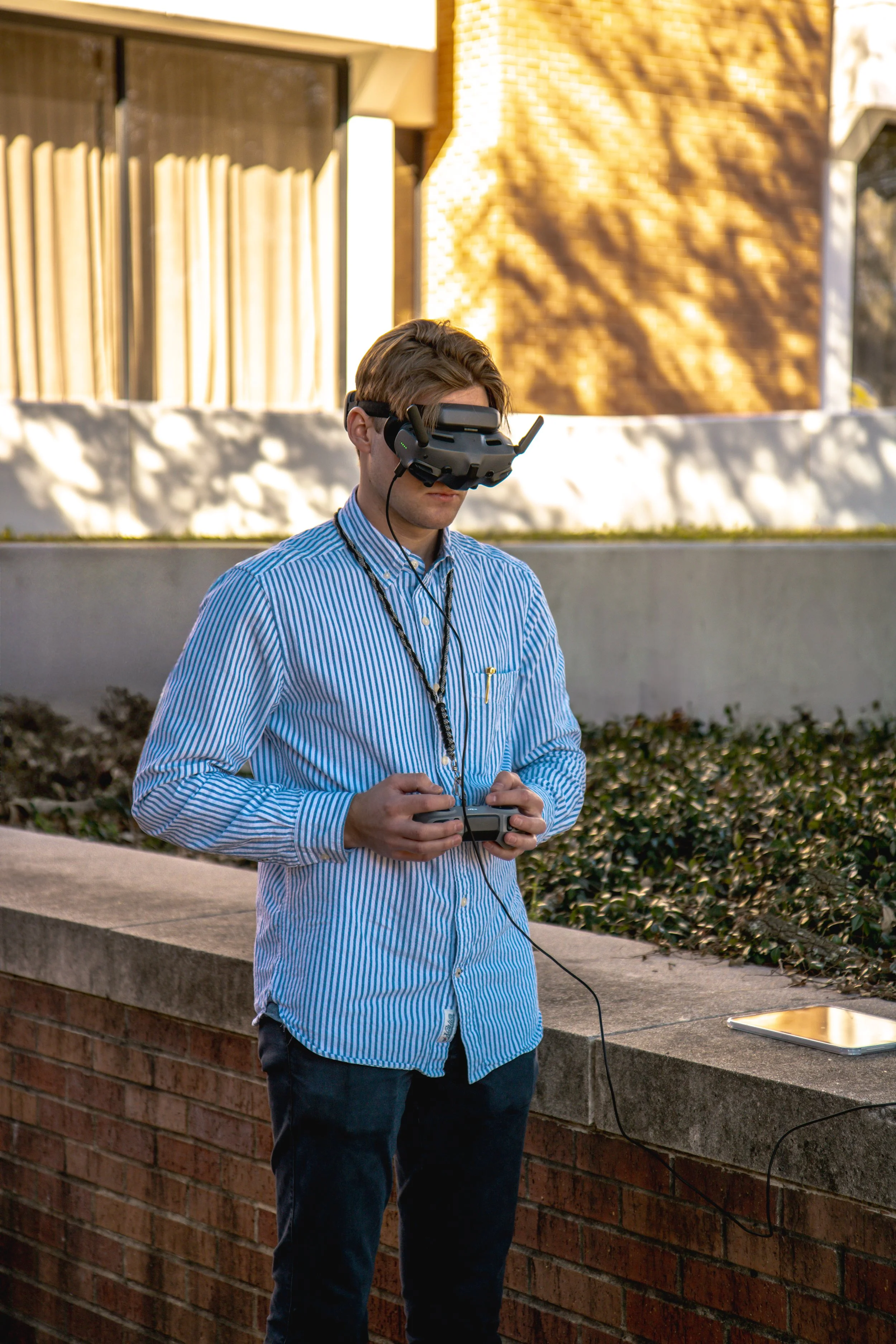 A young man in a blue and white striped shirt using a virtual reality headset outdoors, holding a controller in his hands, with a tablet placed on a brick ledge nearby.