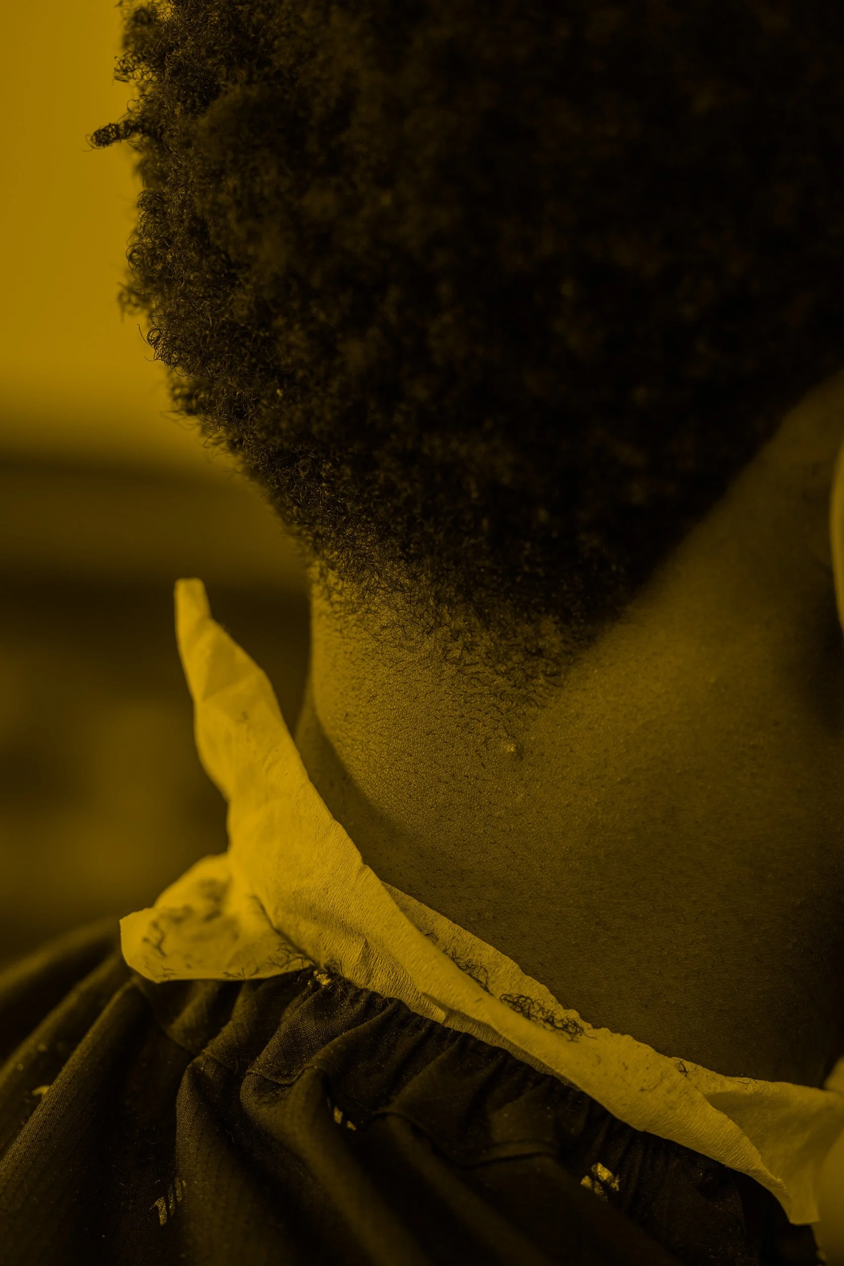 Close-up of a person's neck and chin in warm yellow lighting, showing textured skin and naturally curly dark hair, with a yellow fabric or paper near the shoulder.
