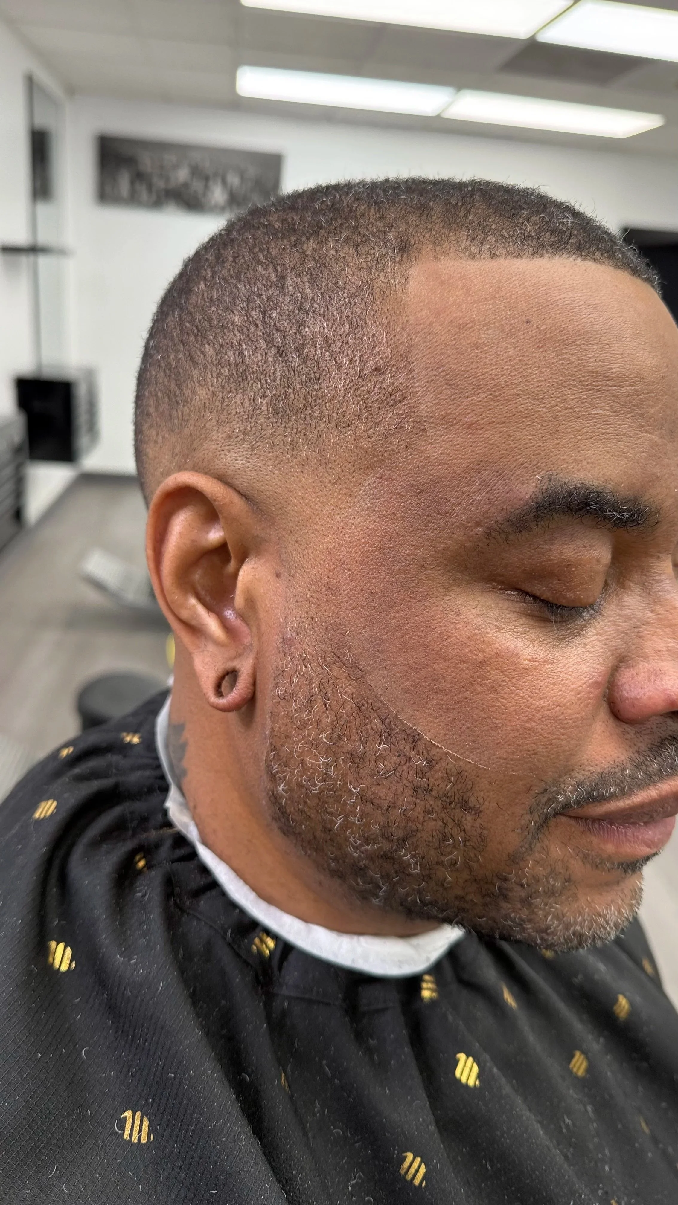 Close-up of a man's face with a fresh buzz cut hairstyle, showing short tapered sides and part of his beard, in a barbershop.