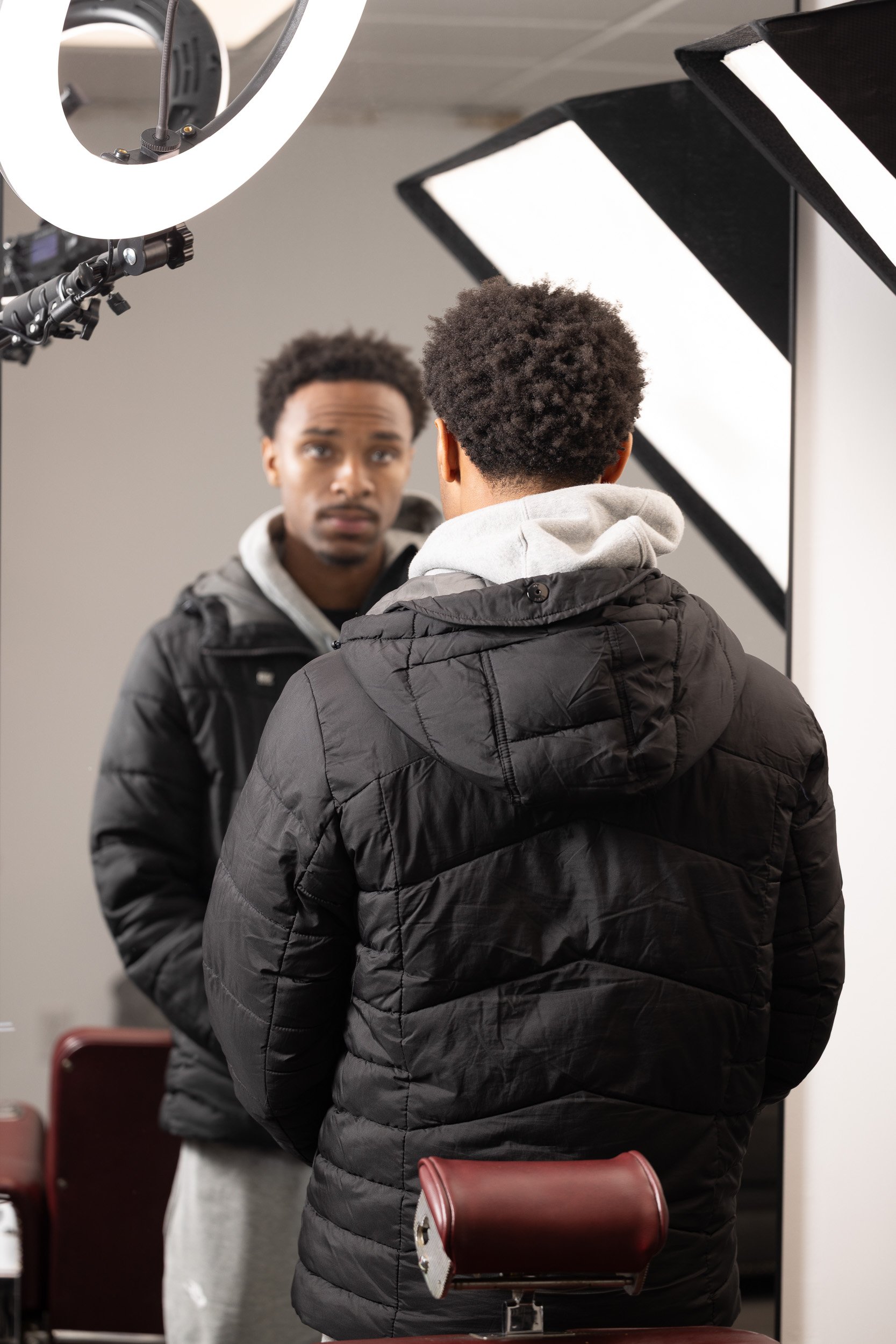 A young man with curly hair wearing a black quilted jacket and gray hoodie, seen from behind, facing a mirror in a photography studio. The studio has professional lighting equipment.