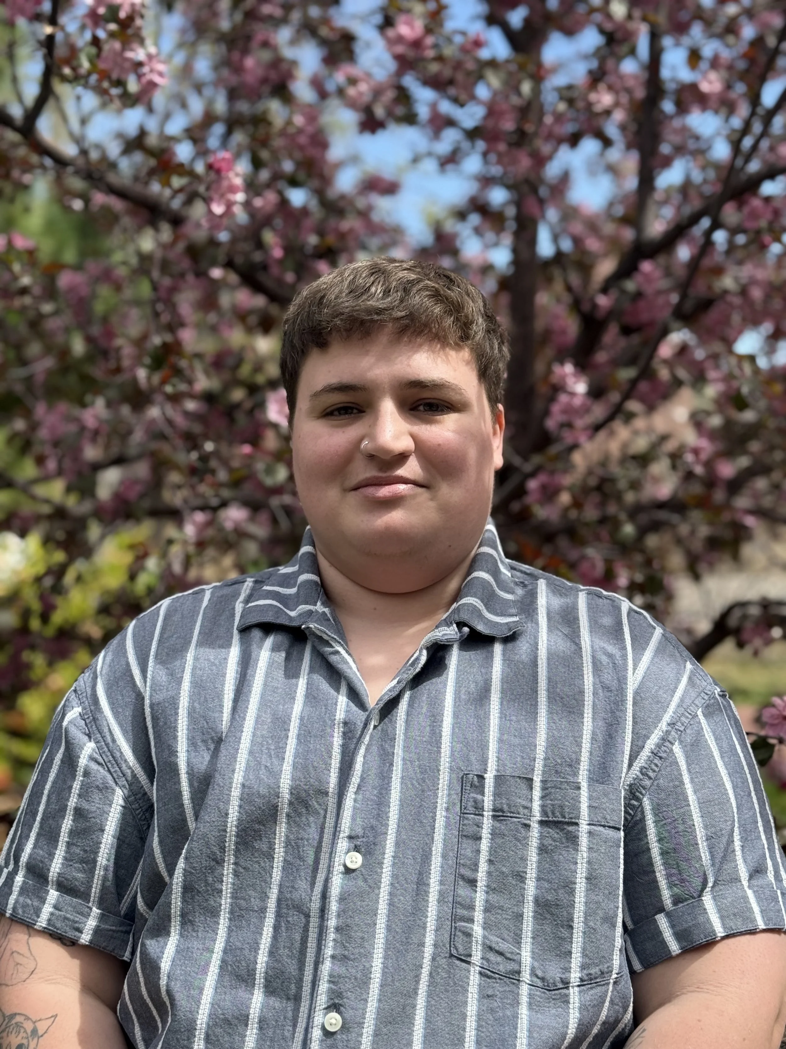 Maveryck headshot in a button up, background of a tree with pink and white flowers