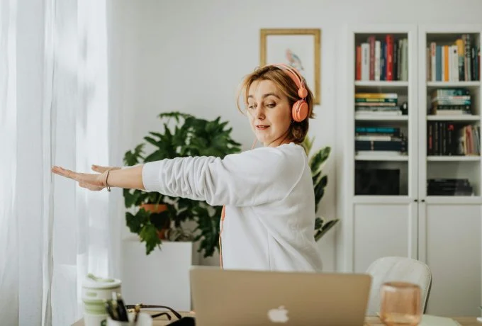 woman in a lovely home office with headphones on stretching in front of laptop