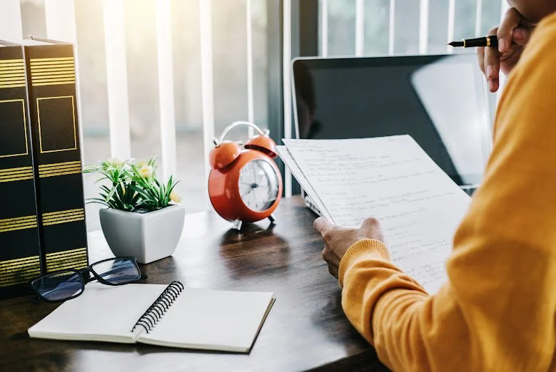 A woman writing notes at a desk with a clock and a notebook