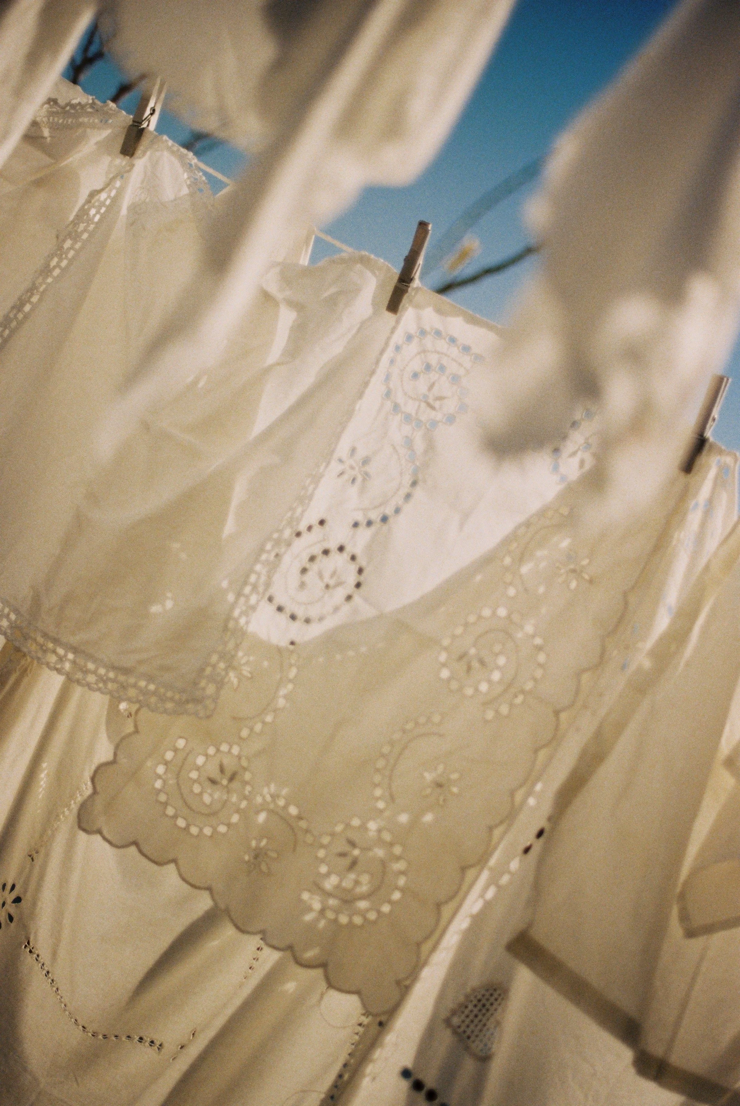 Close-up view of white embroidered fabric hung on a clothesline with clothespins, outdoors with blue sky.