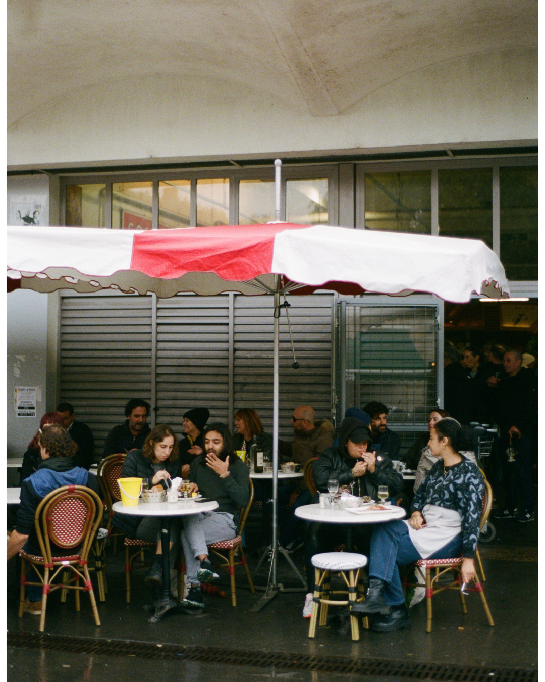People sitting at outdoor tables under a large red and white umbrella at a cafe or restaurant.