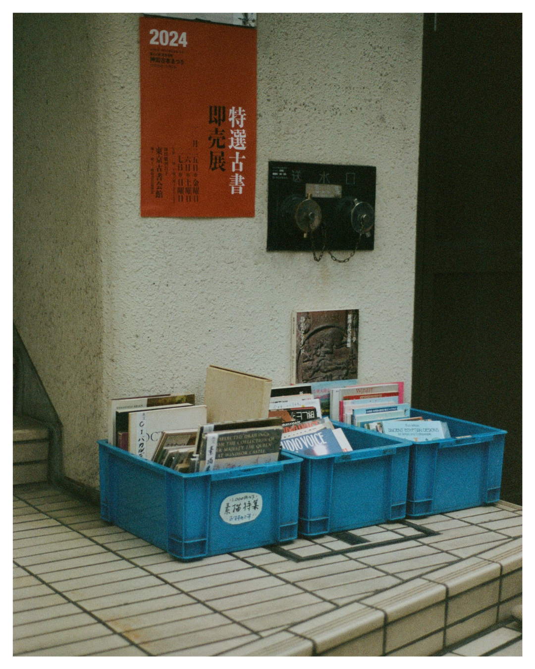 Three blue plastic bins filled with books placed on a tiled surface below a sign with Japanese writing and a black water supply outlet on a beige textured wall.